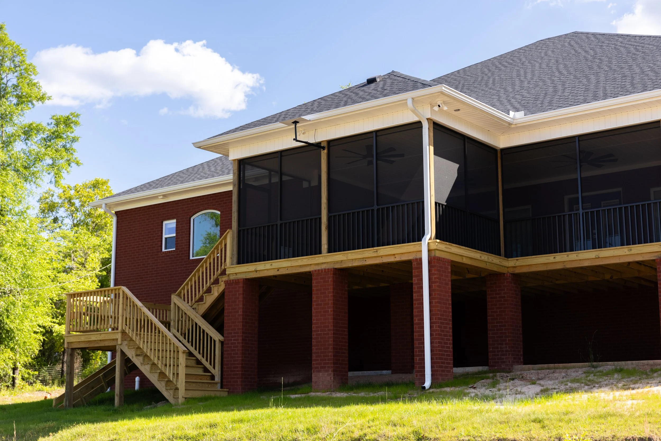 A two-story brick house with a screened-in porch, wooden stairs, and a gray shingled roof in a green backyard with trees and blue sky.