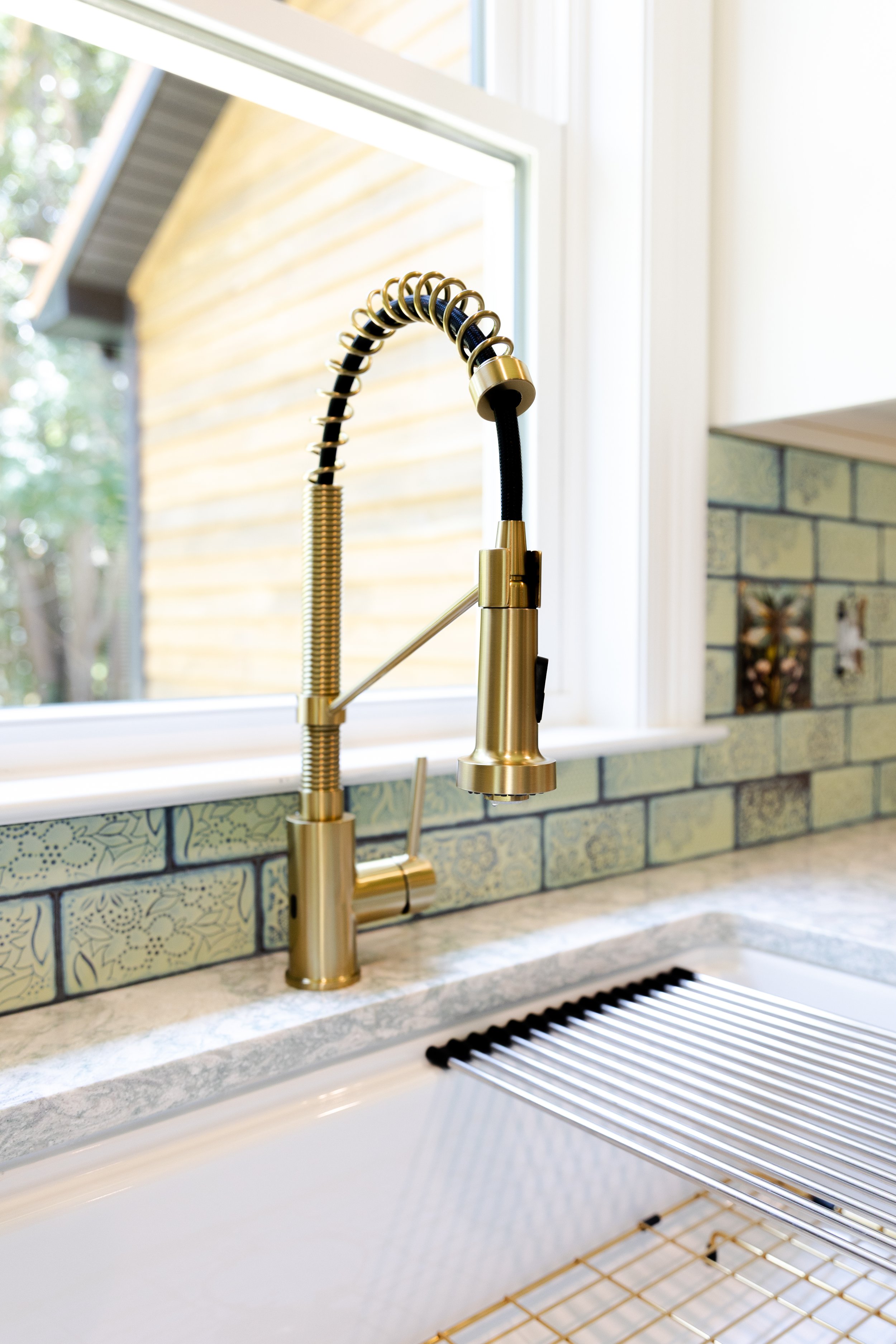 Close-up of a modern gold kitchen sink faucet with a pull-down sprayer, situated under a window with a view of a yellow house's exterior and a tree with green leaves.
