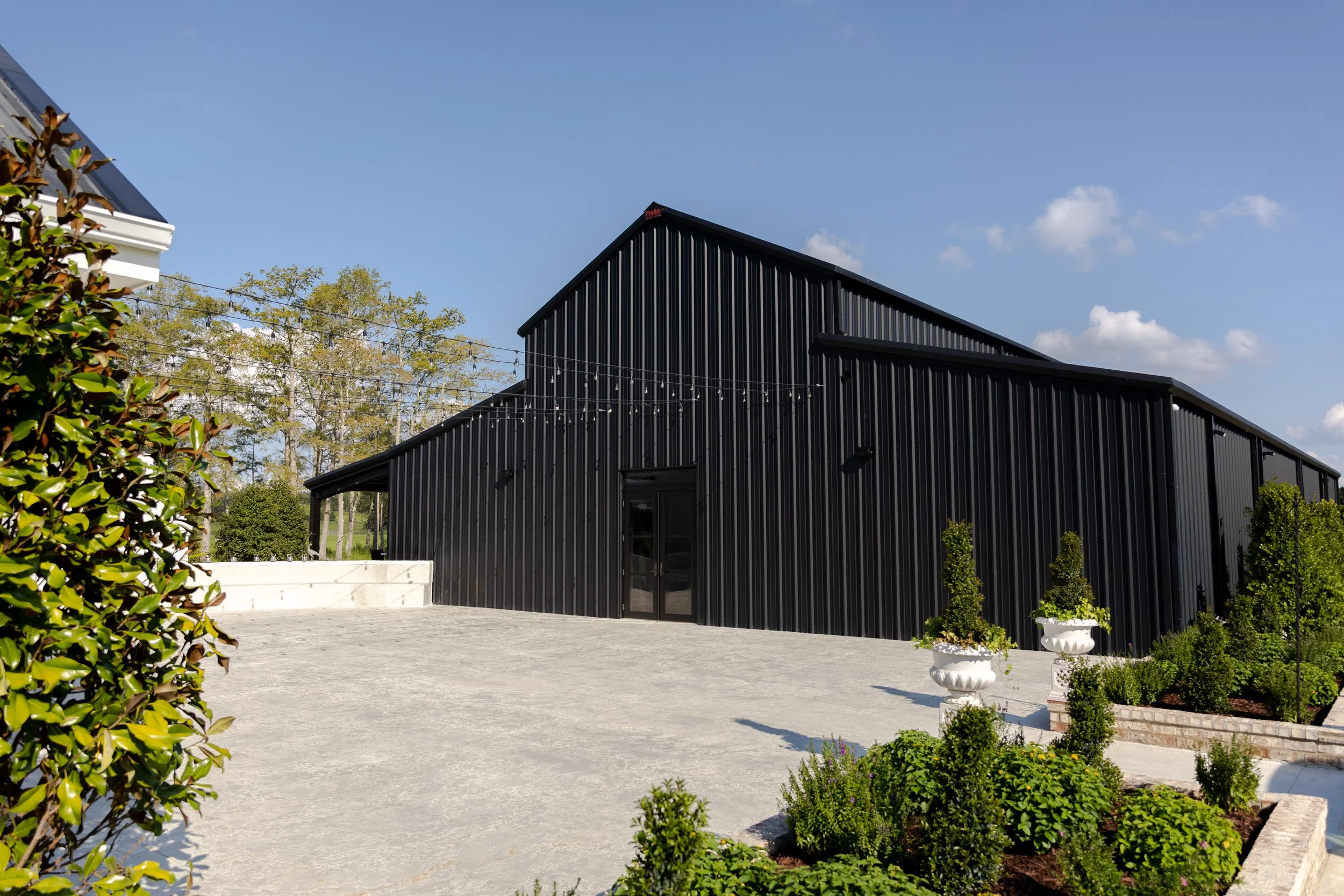 A modern black metal building with a sloped roof, surrounded by greenery and potted plants, under a clear blue sky.