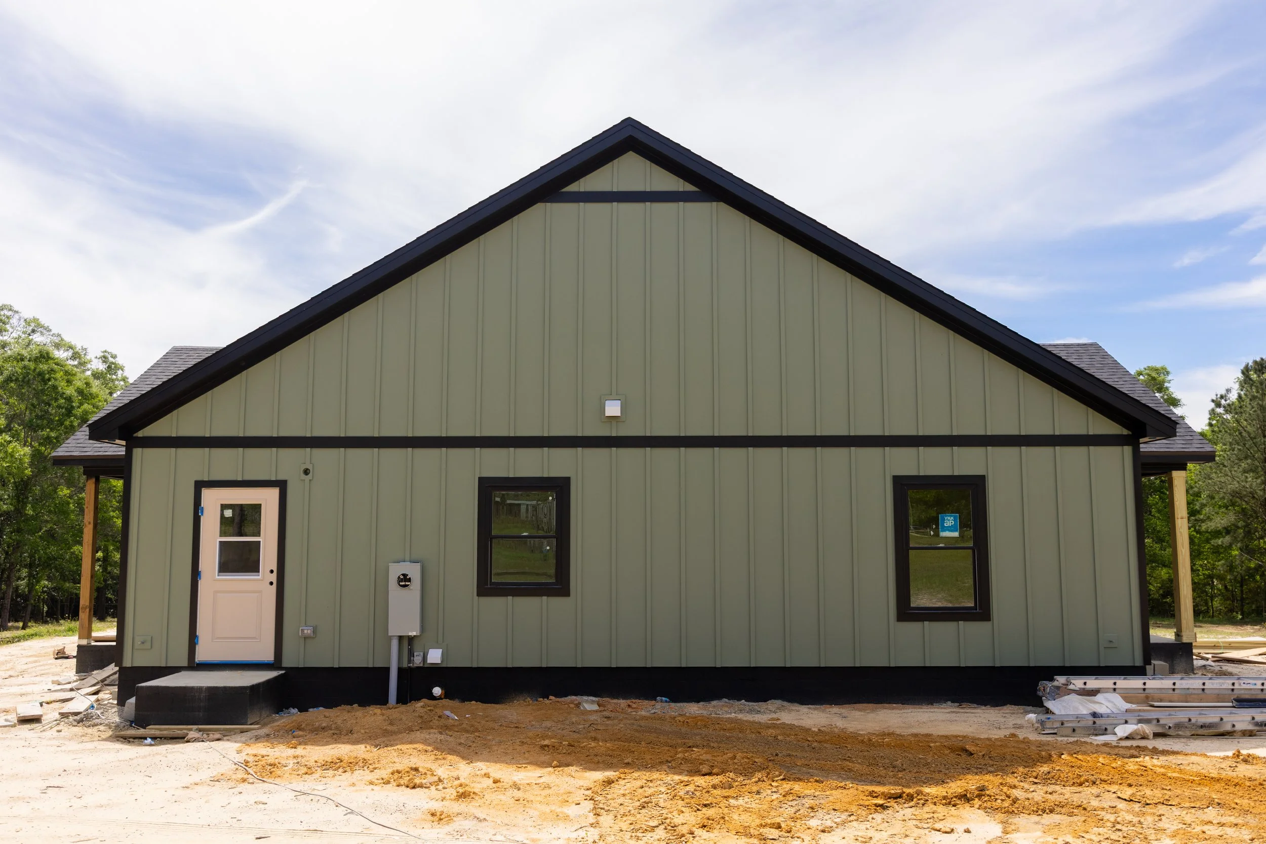 New house under construction with pale yellow exterior siding, black window and door trim, and a black roof against a partly cloudy sky, with construction materials and dirt ground in the foreground.