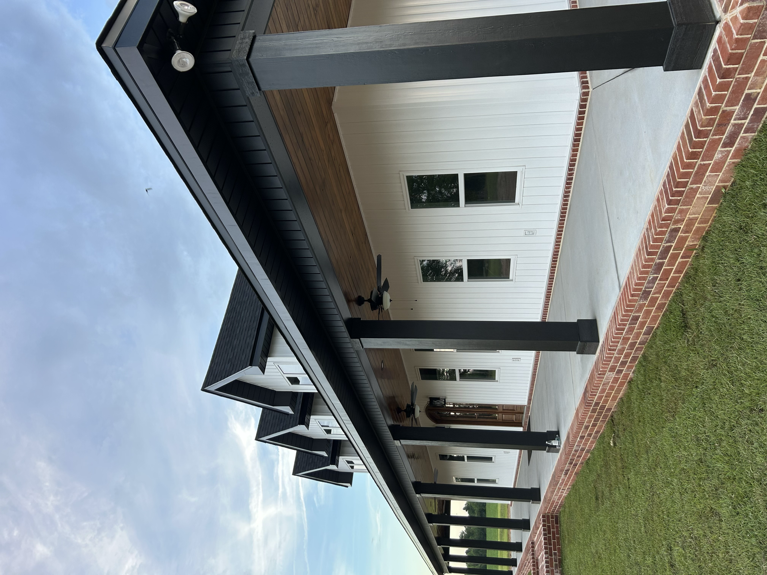 A modern house exterior with black roof and trim, beige siding, large windows, a brick foundation, and a concrete patio.