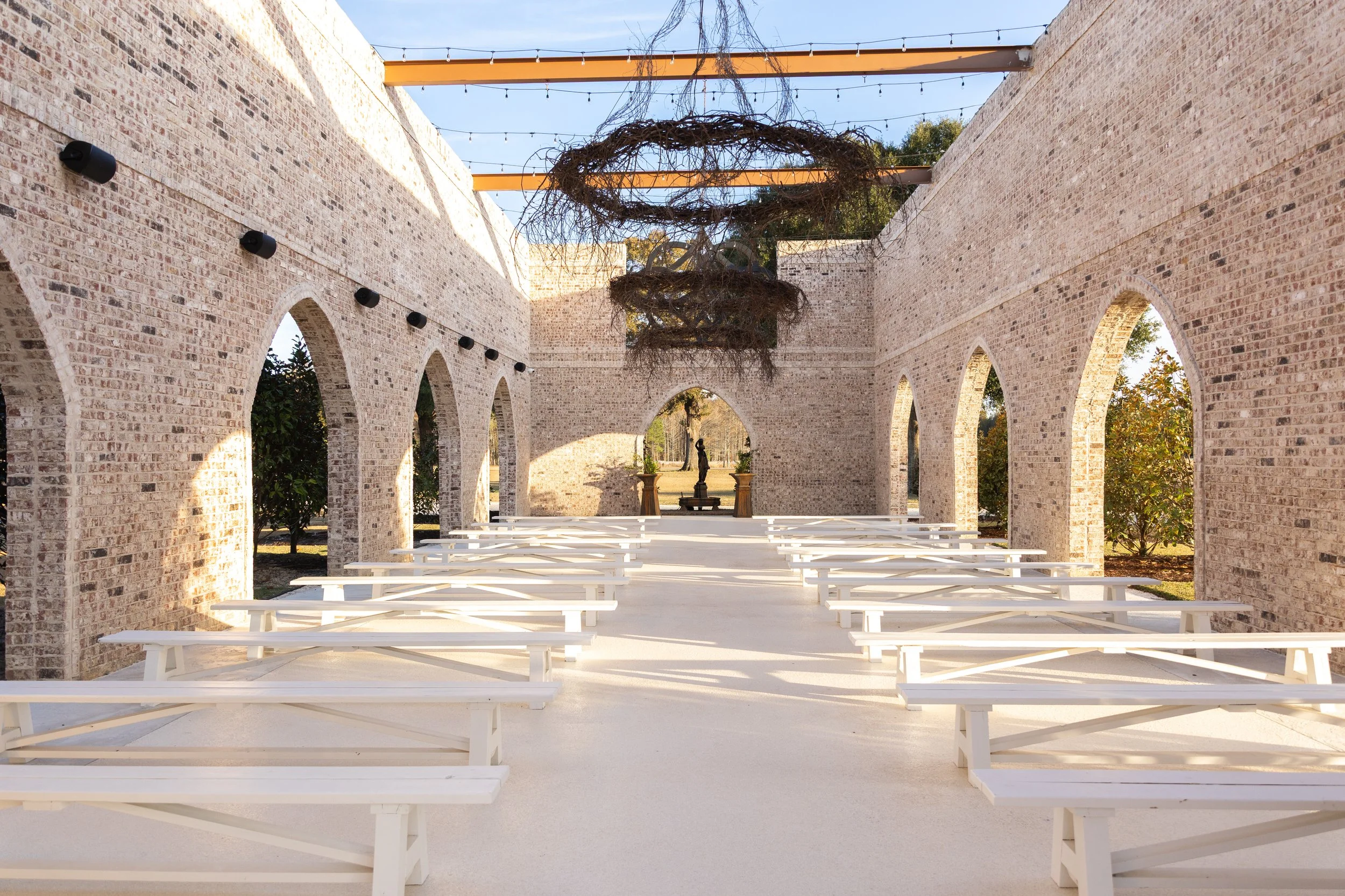 An outdoor wedding ceremony setup with white wooden benches arranged in rows on a light-colored concrete floor inside a brick archway structure. The structure has a series of brick arches and black wall-mounted lights. Above, there are string lights 
