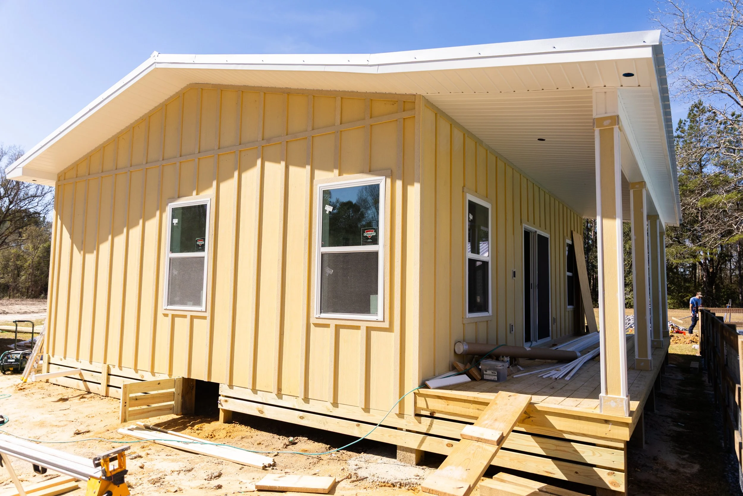 View of a house under construction with yellow exterior siding, multiple windows, and a large overhanging roof, surrounded by a dirt area and trees in the background.