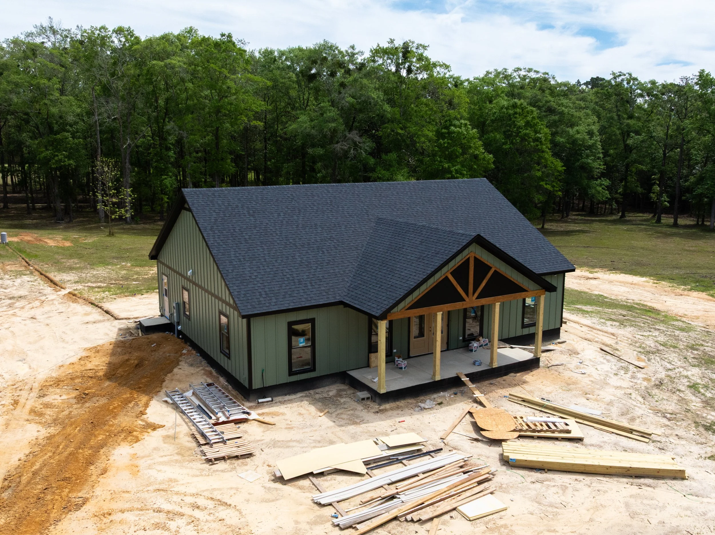 A house under construction in a partially wooded area with a dirt ground and building materials scattered around.