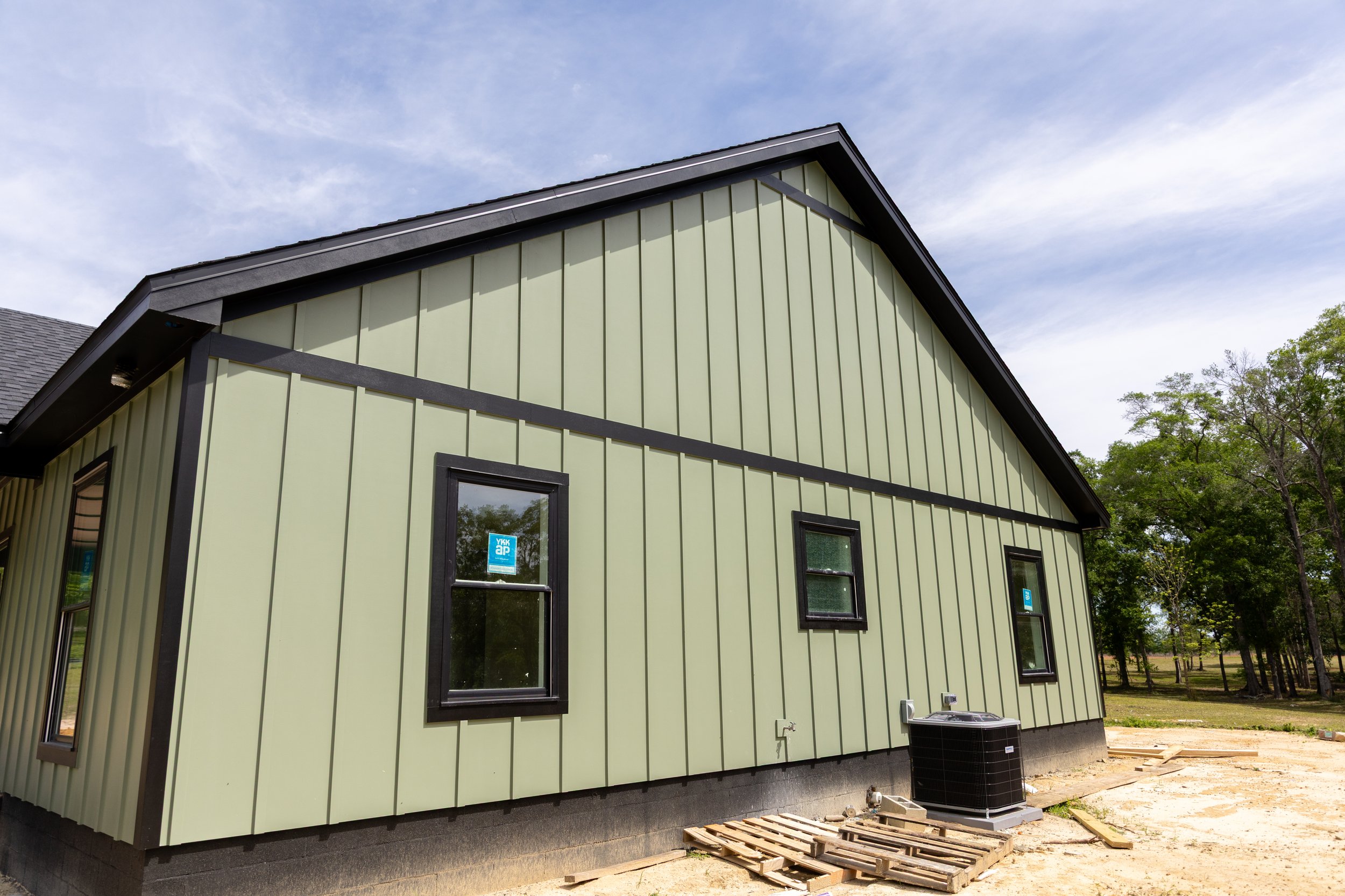 Front of a new house with light green vertical siding and black trim, several windows, and an air conditioning unit outside, under a partly cloudy sky.