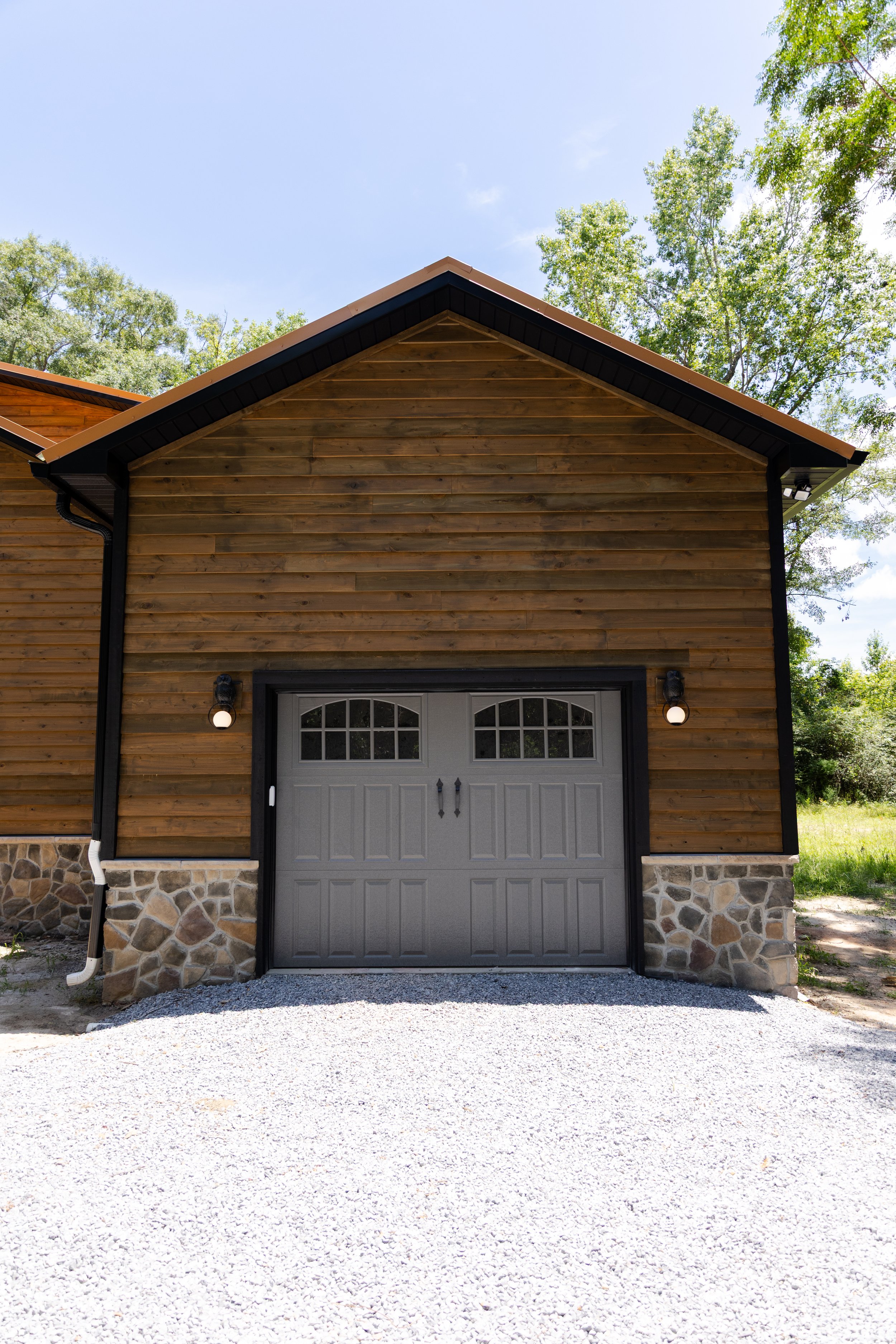 A garage with wooden siding, a stone foundation, and a gray garage door with windows at the top, set against a background of green trees and a blue sky.