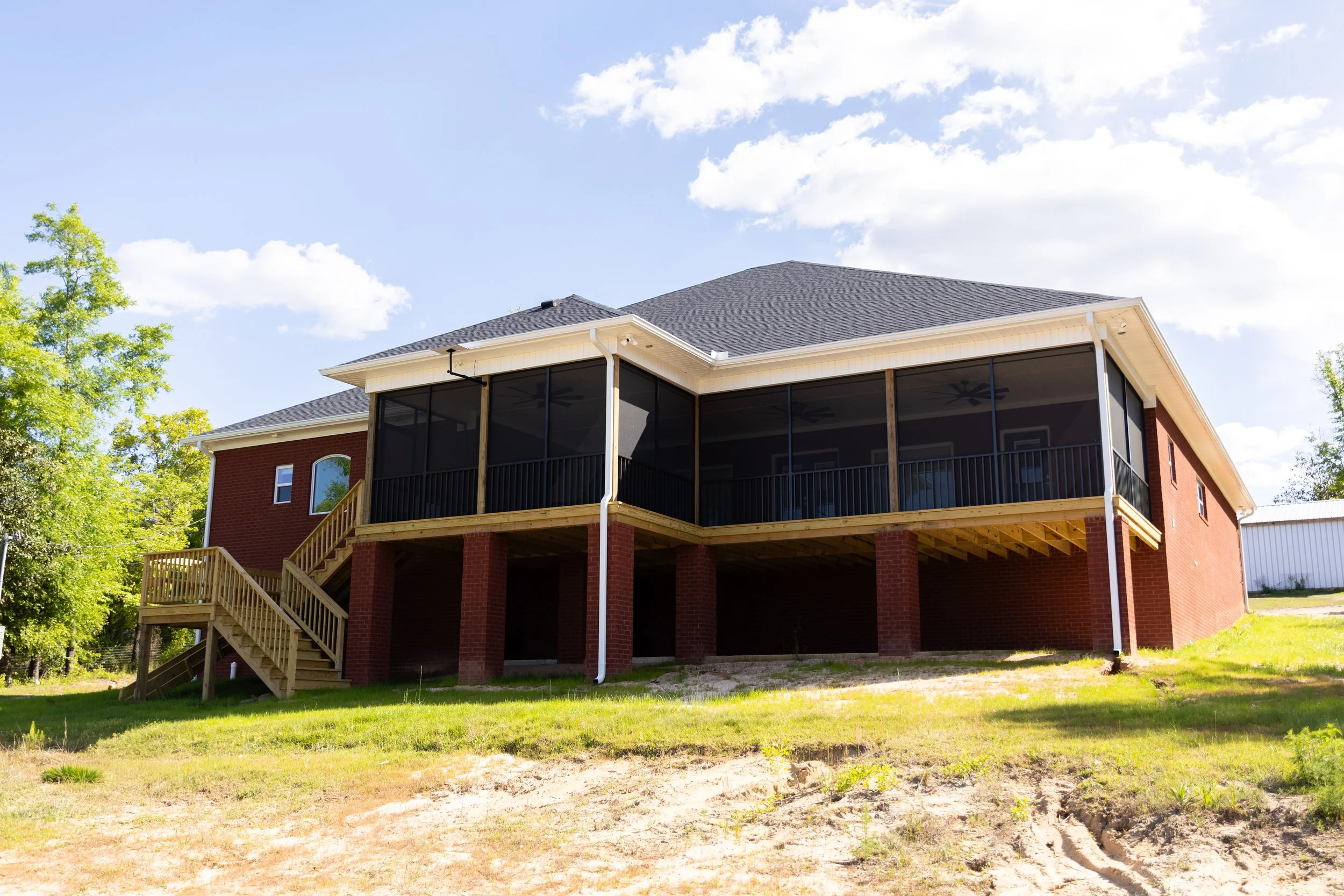 The back of a house with a screened porch, brick exterior, wooden stairs, and a gable roof, with trees and a blue sky in the background.