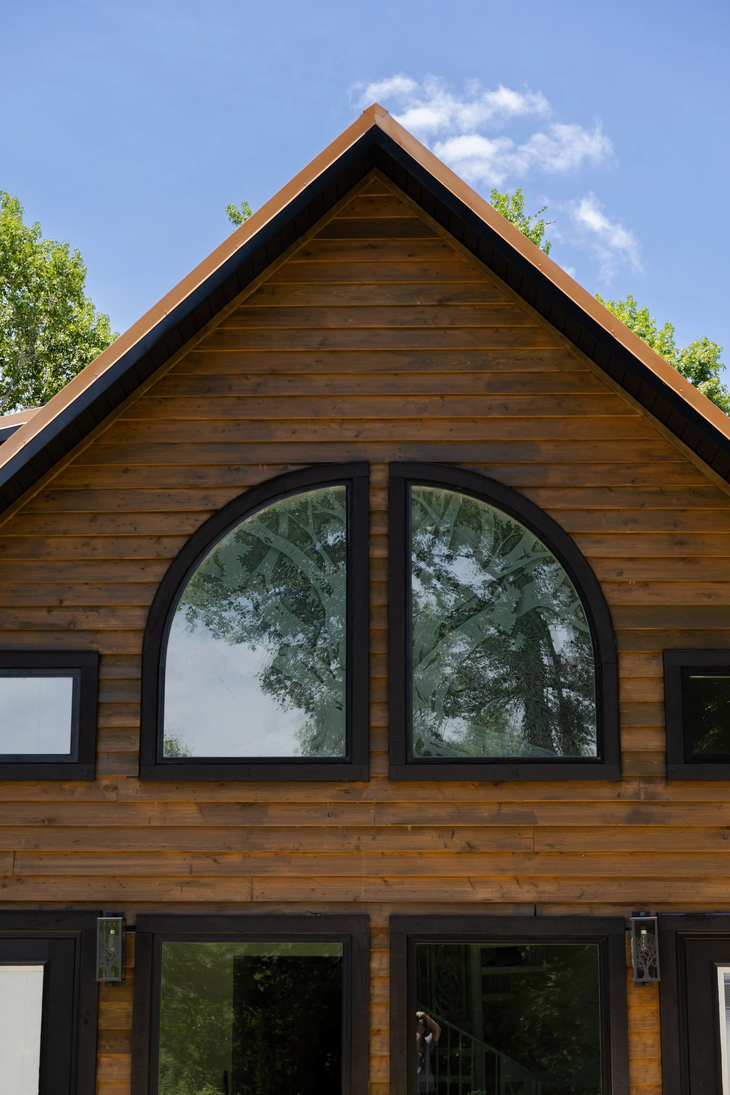 Front view of a wooden house with large, arched windows and a steeply pitched roof, surrounded by trees on a sunny day with a few clouds in the blue sky.