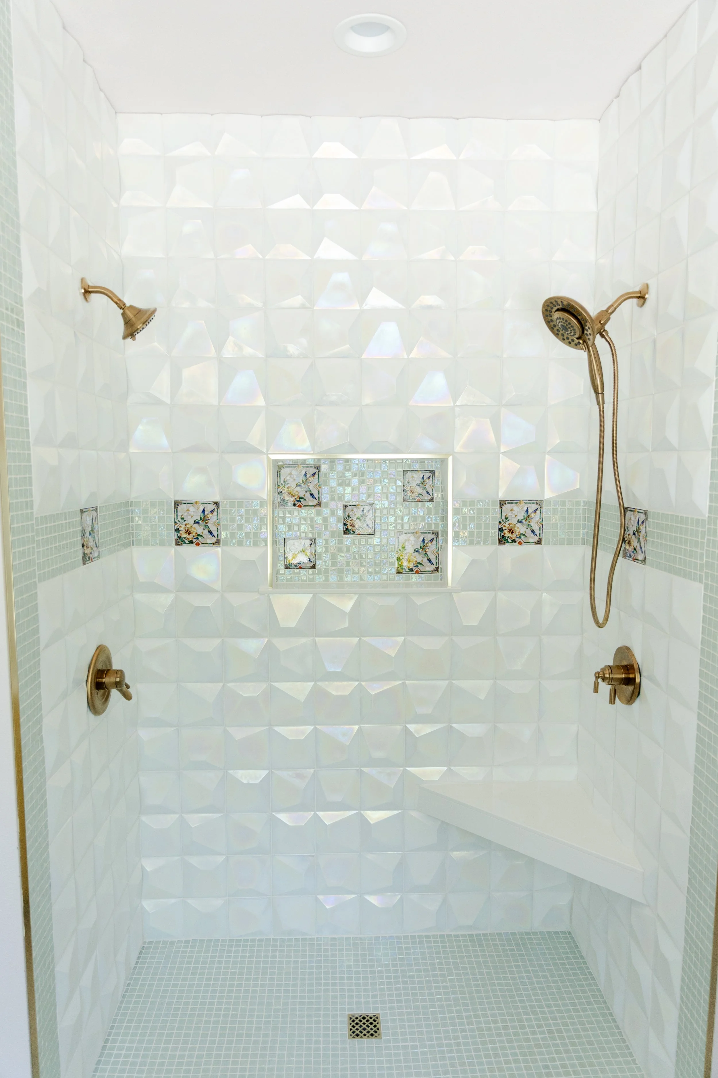 A spacious walk-in shower with white 3D tiled walls, decorative mosaic tiles featuring birds and flowers near the middle, a brushed bronze manual shower head and a handheld shower head. The floor is covered in small light gray tiles with a drain in t