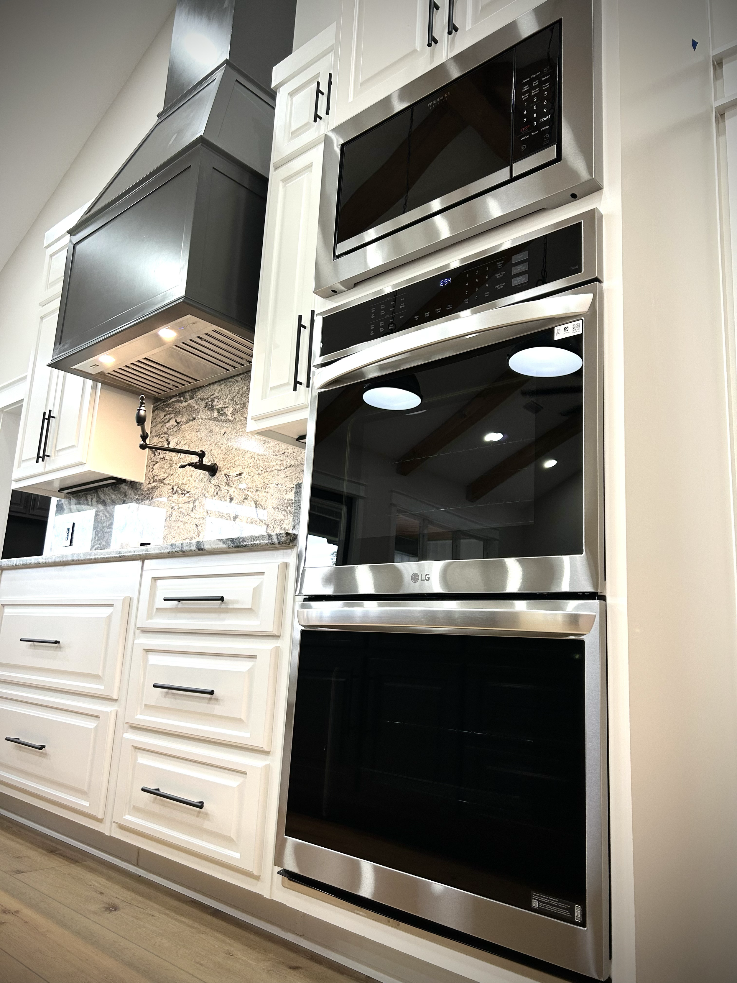 A close-up view of modern kitchen appliances, including a built-in microwave oven and a double oven, installed within white kitchen cabinets with black handles. The kitchen features a granite countertop and a wood floor.