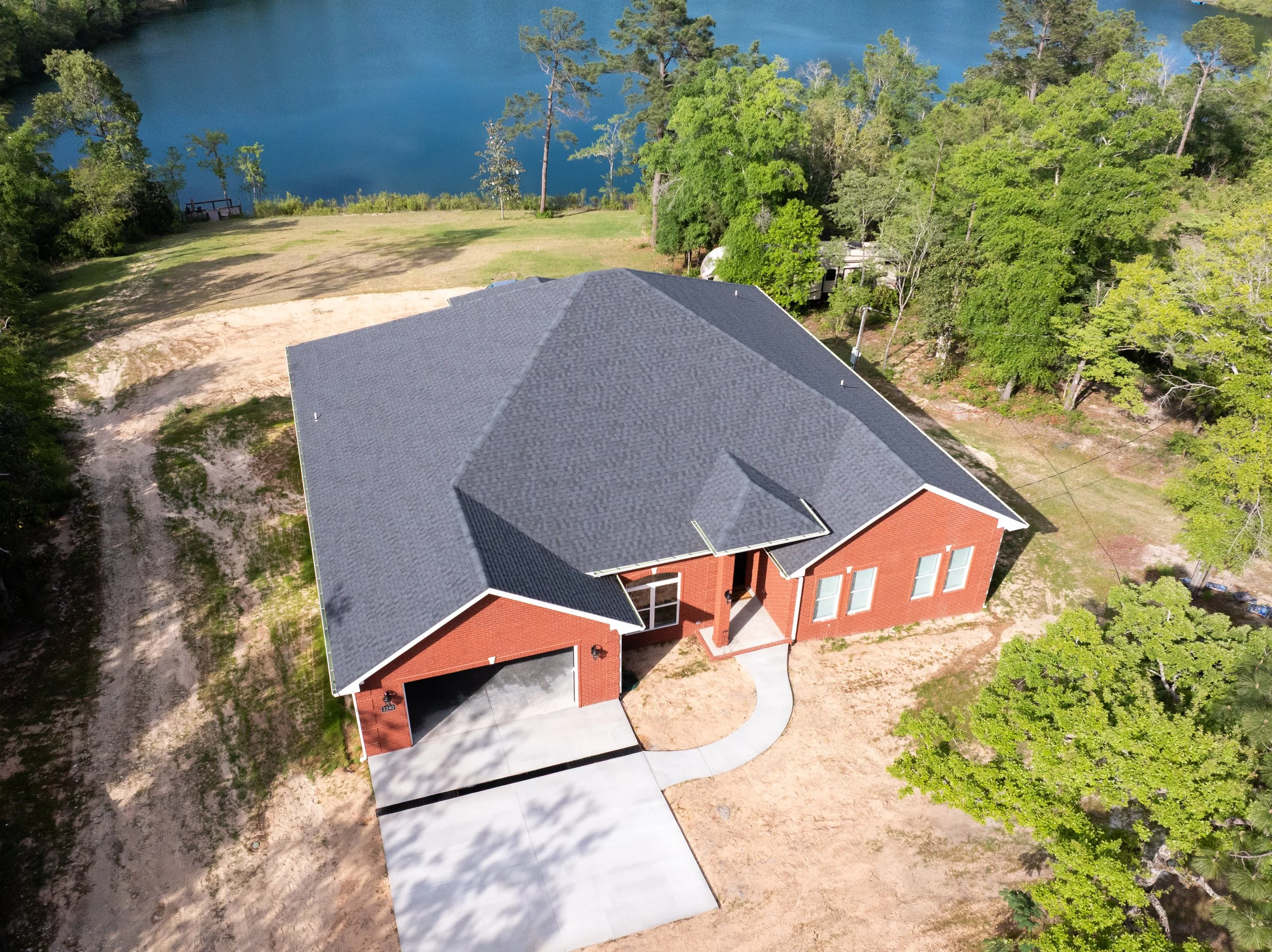 Aerial view of a red brick house with gray shingled roof, driveway, surrounded by green trees, near a body of water.