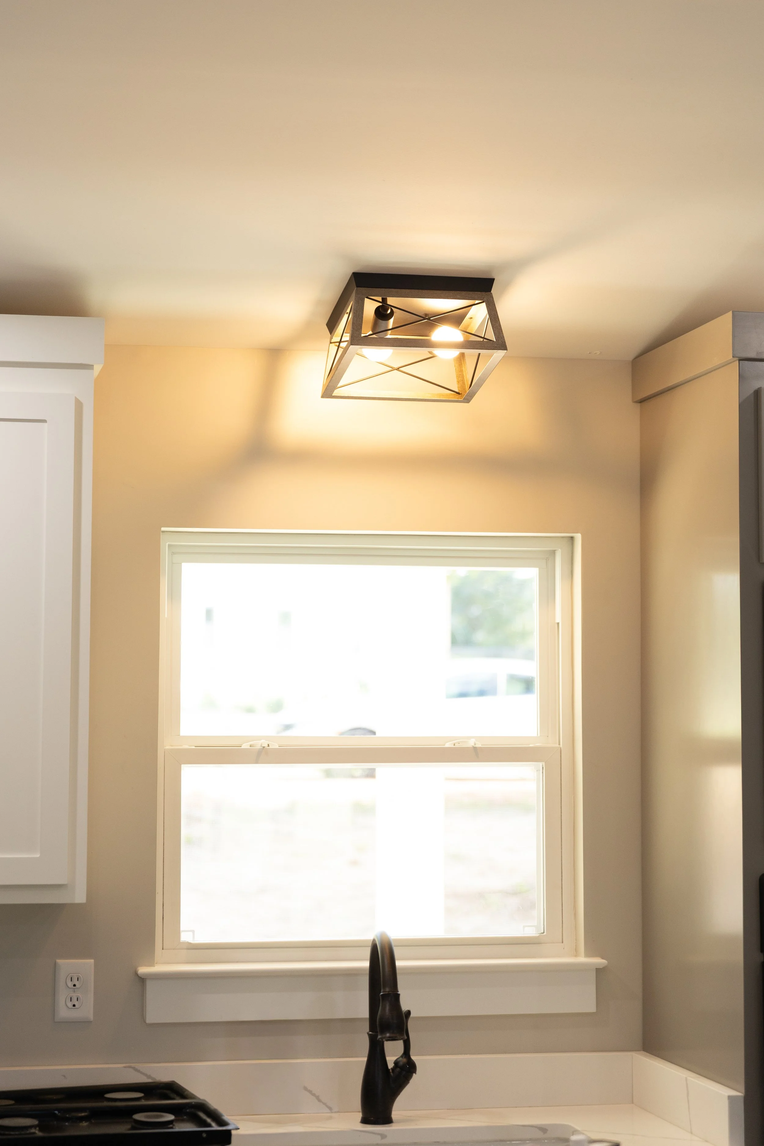 Kitchen window above sink with a black faucet, white frame, beige walls, and a ceiling light fixture.