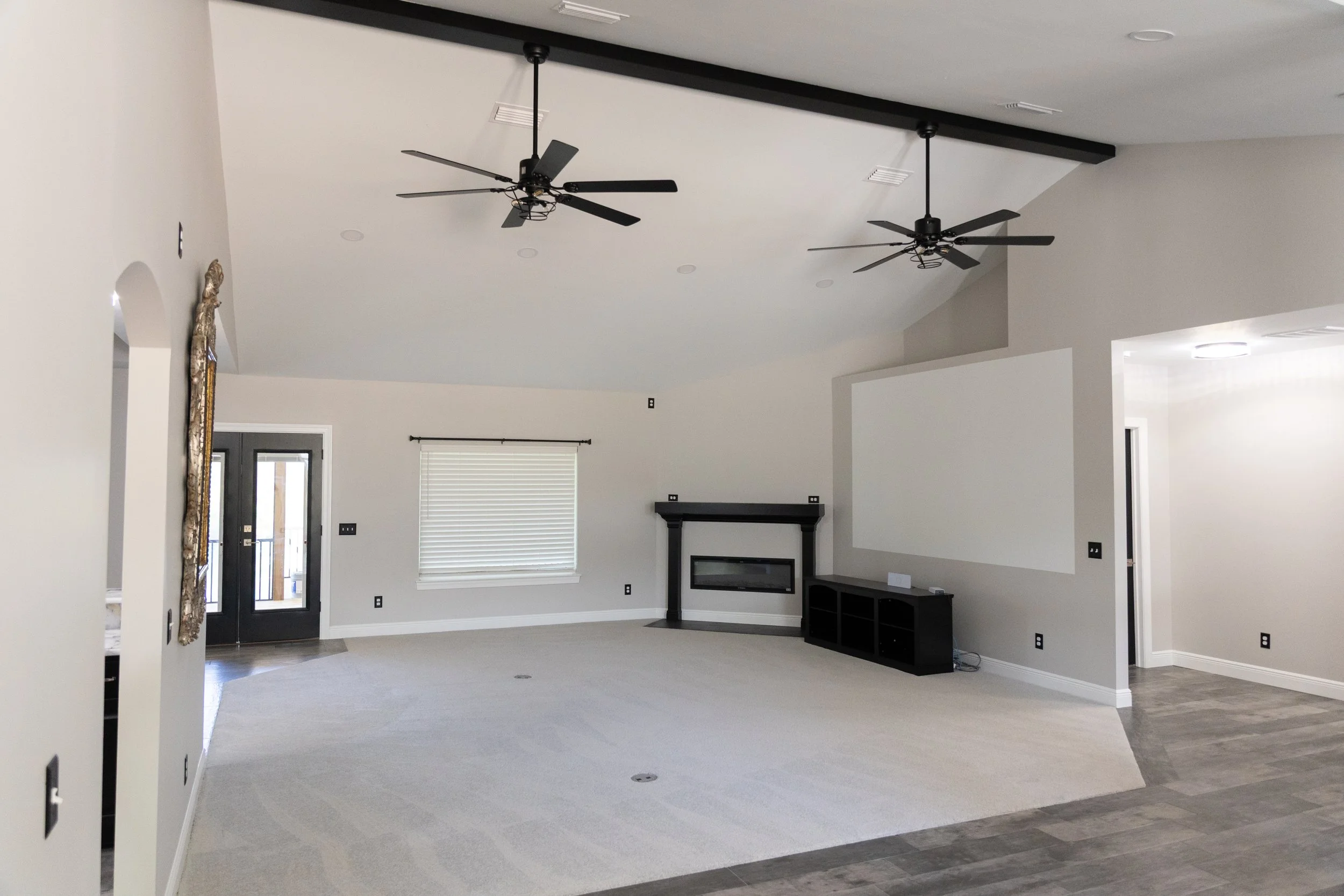 Empty living room with white walls, beige carpet, two black ceiling fans, a fireplace with a black mantel, and large window with closed white blinds.