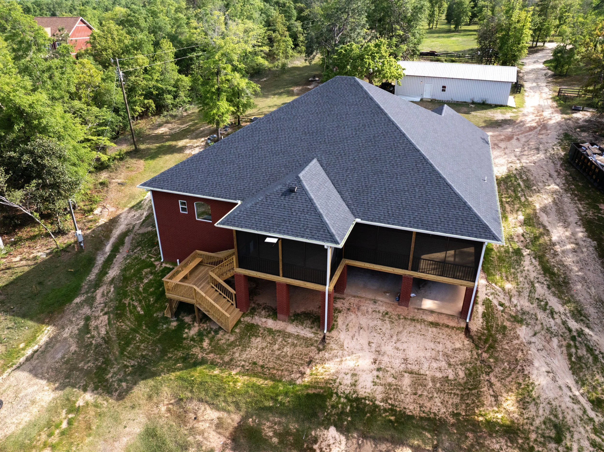 Aerial view of a house with a dark gray shingled roof and red brick walls, surrounded by green trees, a white shed, and a dirt driveway.