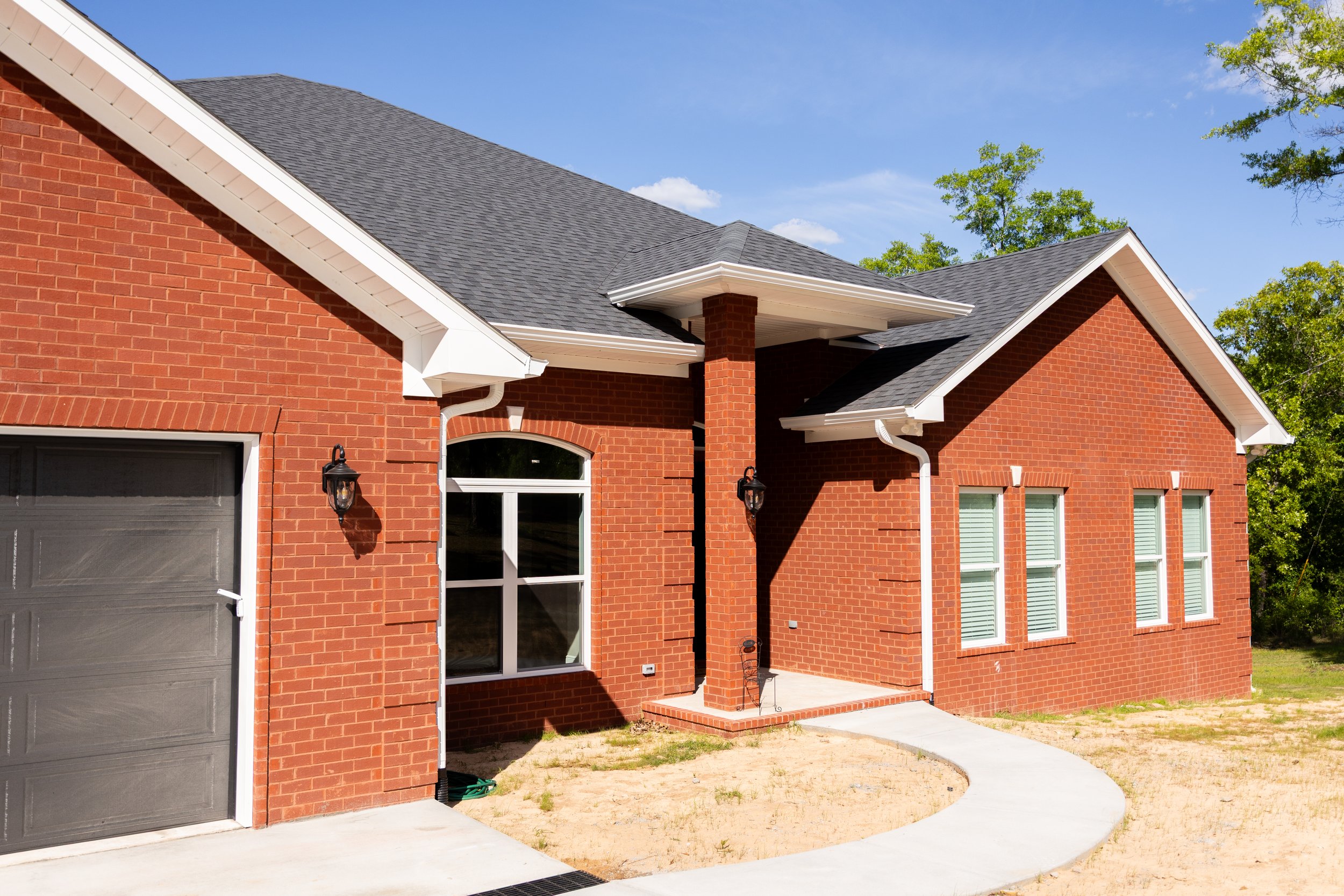 A brick house with a gray door, large windows, a garage, a small front porch with brick columns, a curved concrete walkway, and a yard with sparse grass and dirt, surrounded by green trees under a bright blue sky.