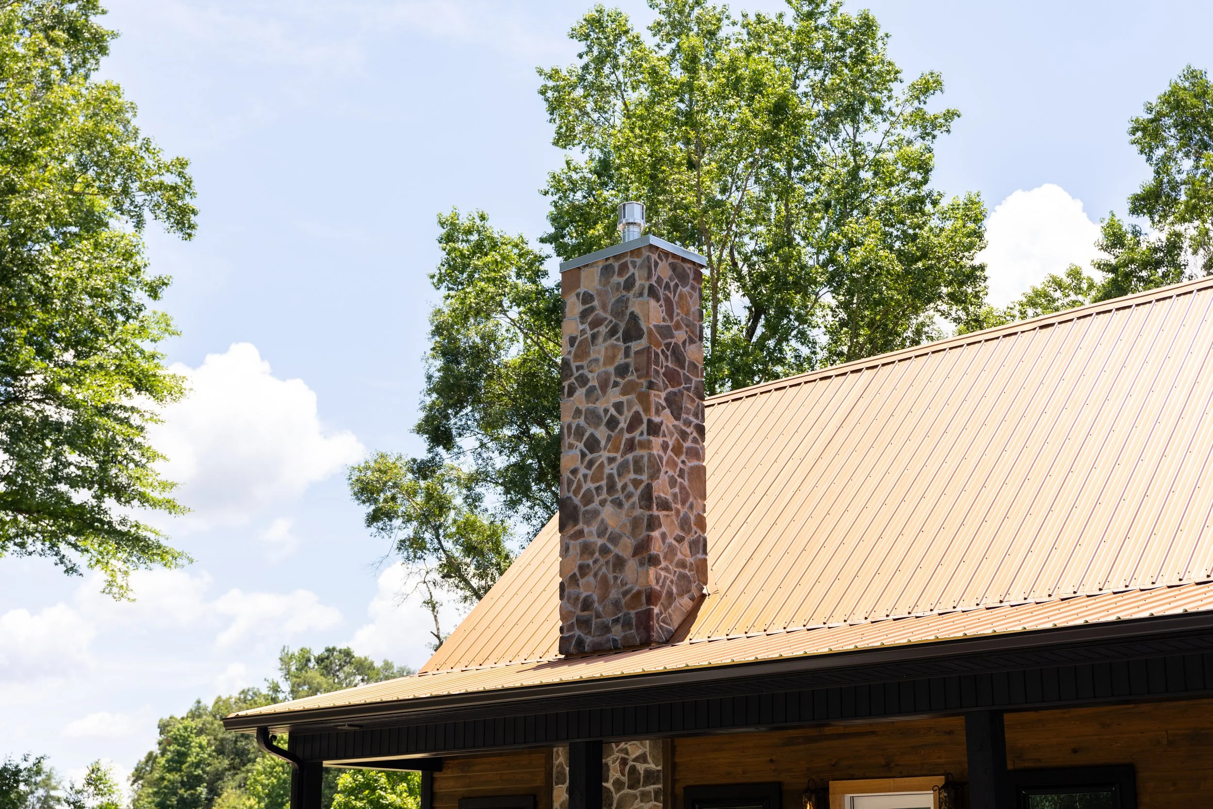 A house roof with a stone chimney and a metal cap, surrounded by tall green trees under a partly cloudy sky.