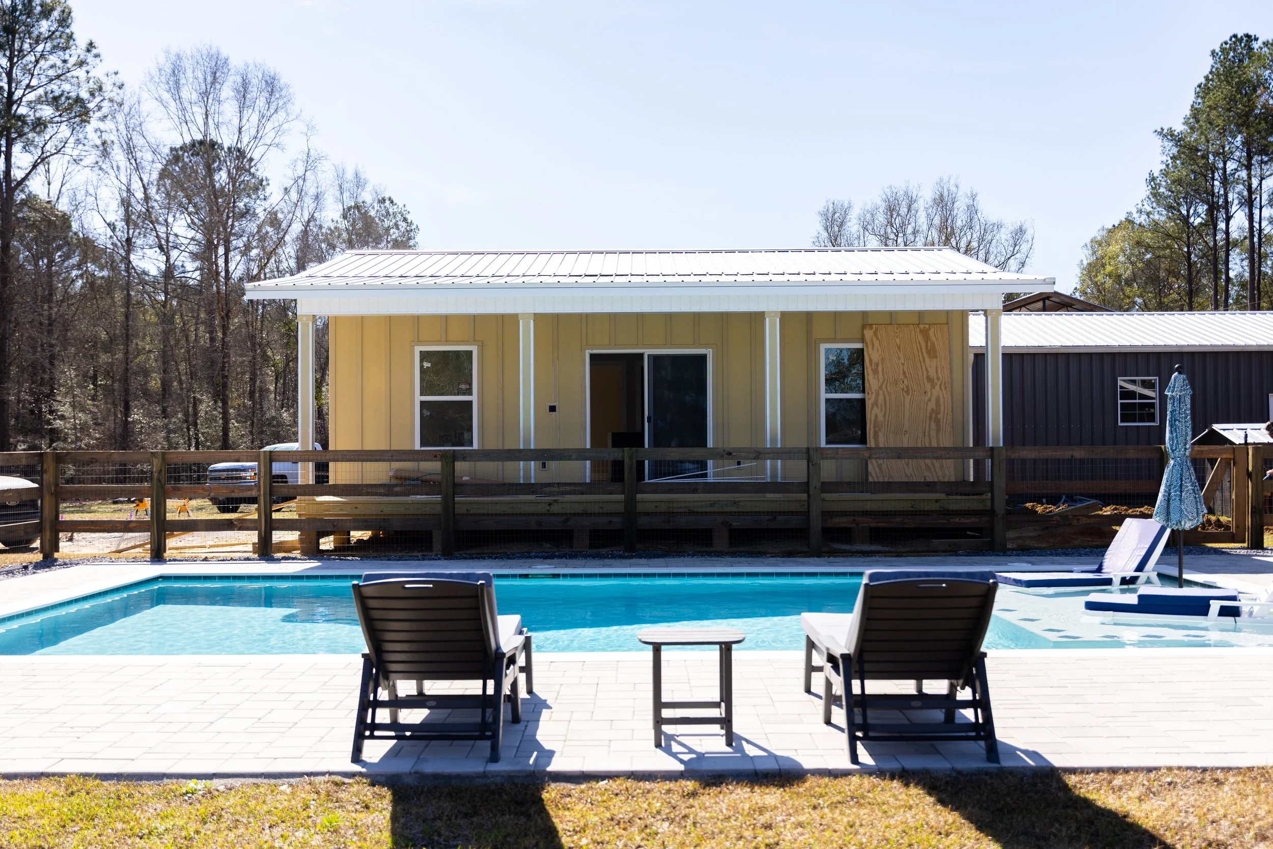 Backyard with swimming pool, poolside chairs, wooden fence, small house or shed with yellow siding, and trees in the background.