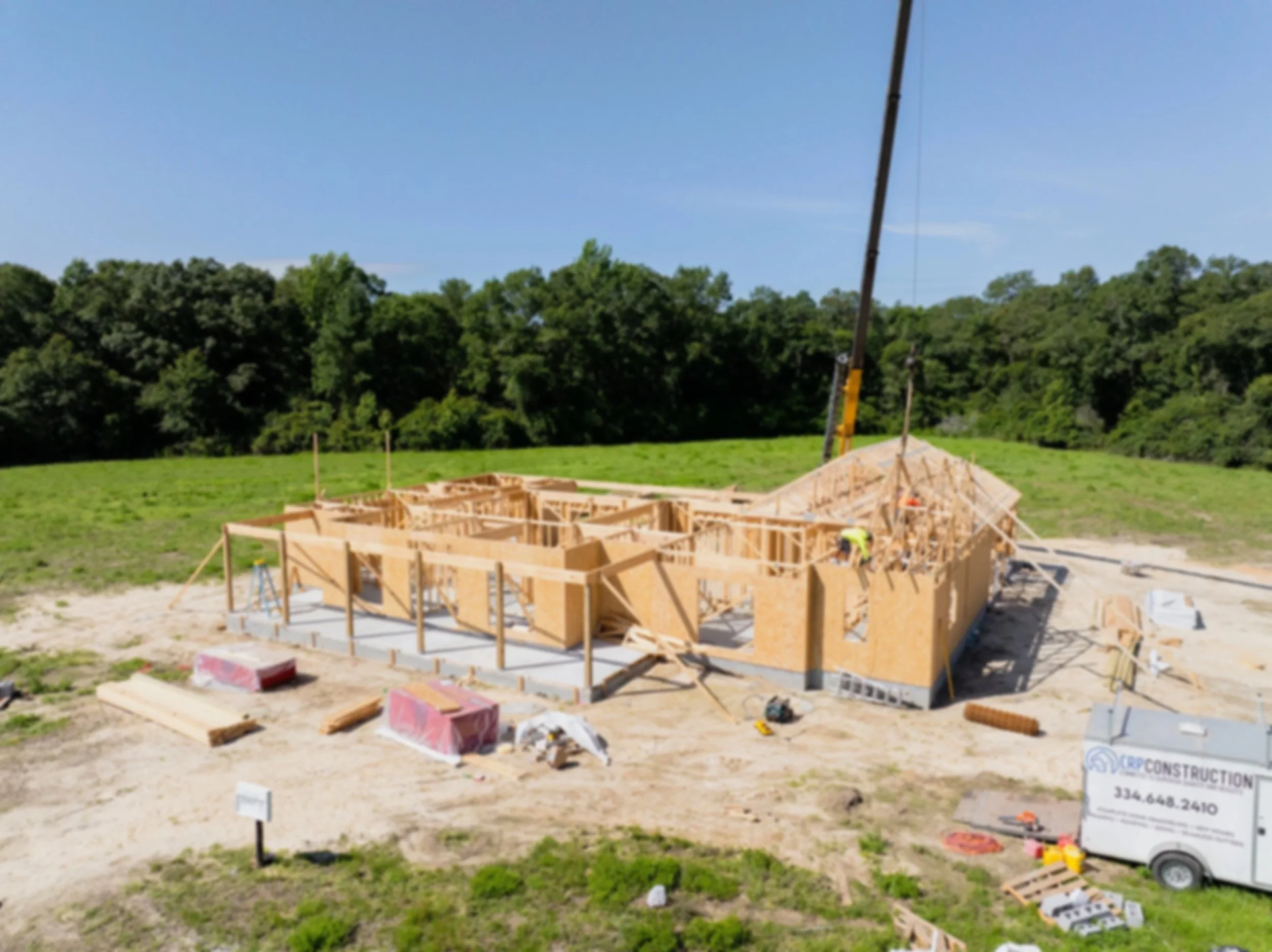 Construction site with wooden framework of a building, construction workers, and a crane in the background, surrounded by greenery and trees.