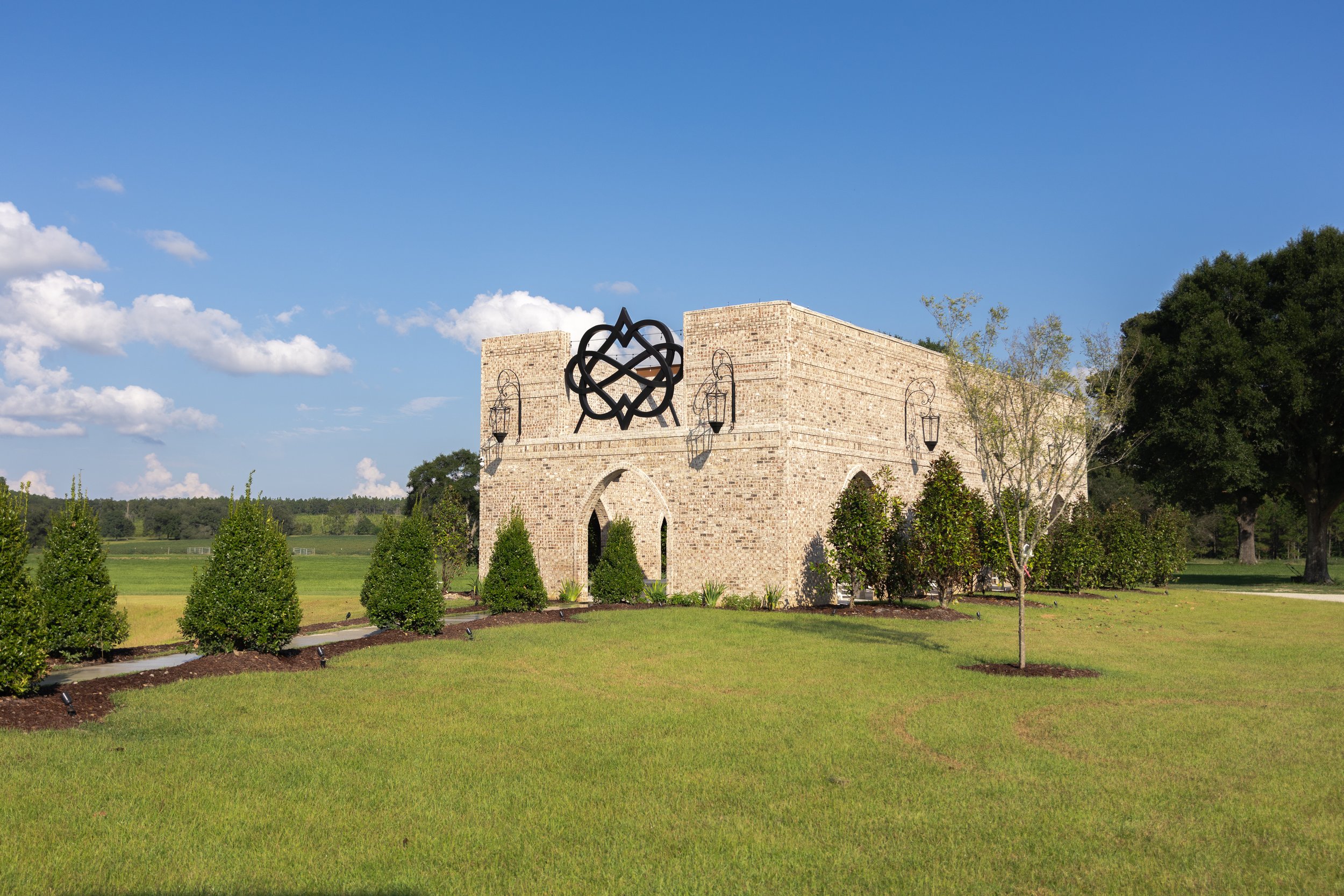 A brick structure with a decorative metal sculpture on top, surrounded by neatly trimmed bushes and trees under a bright blue sky with scattered clouds.