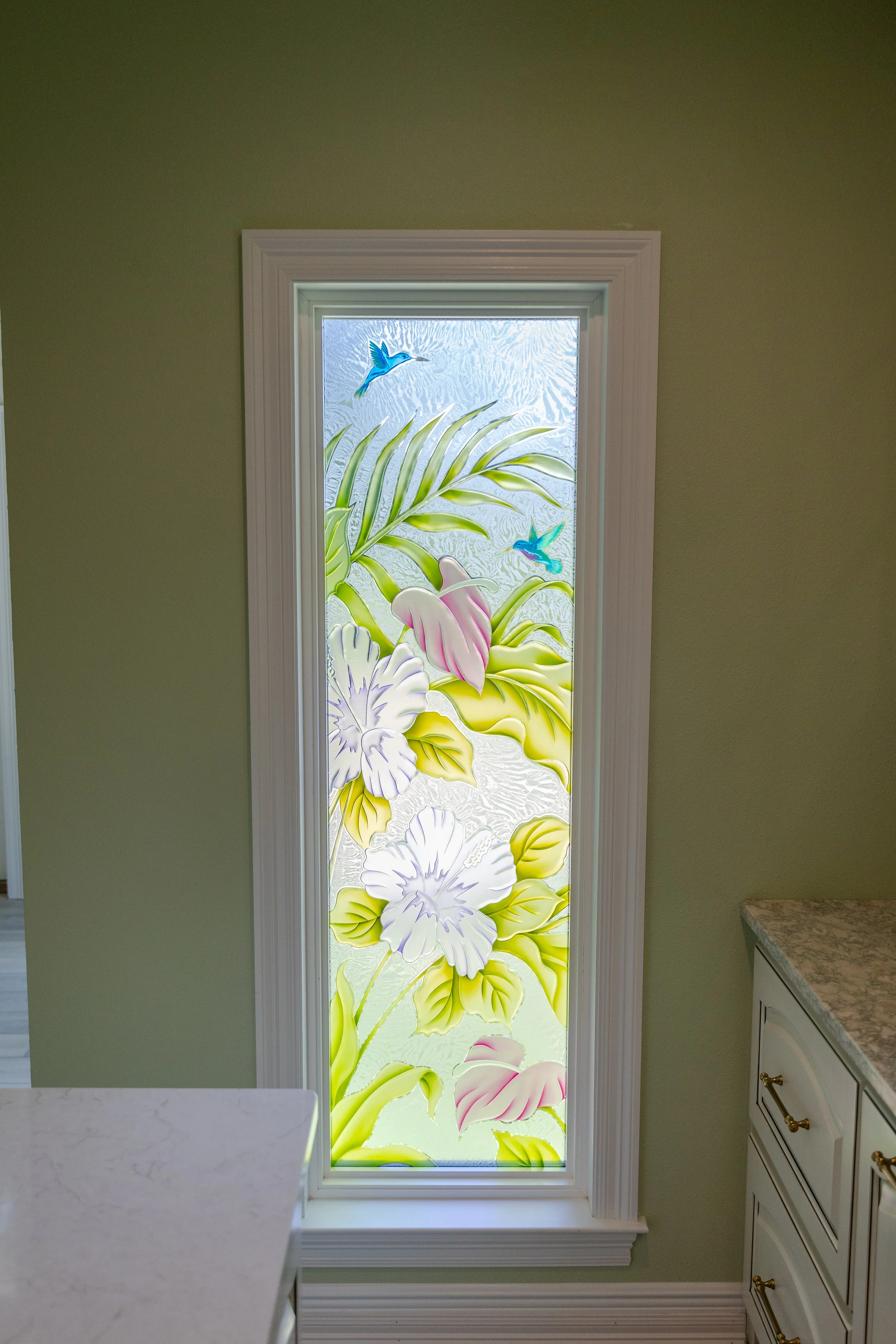 A decorative glass door with a tropical floral and bird design, in a kitchen with green walls and white cabinets.