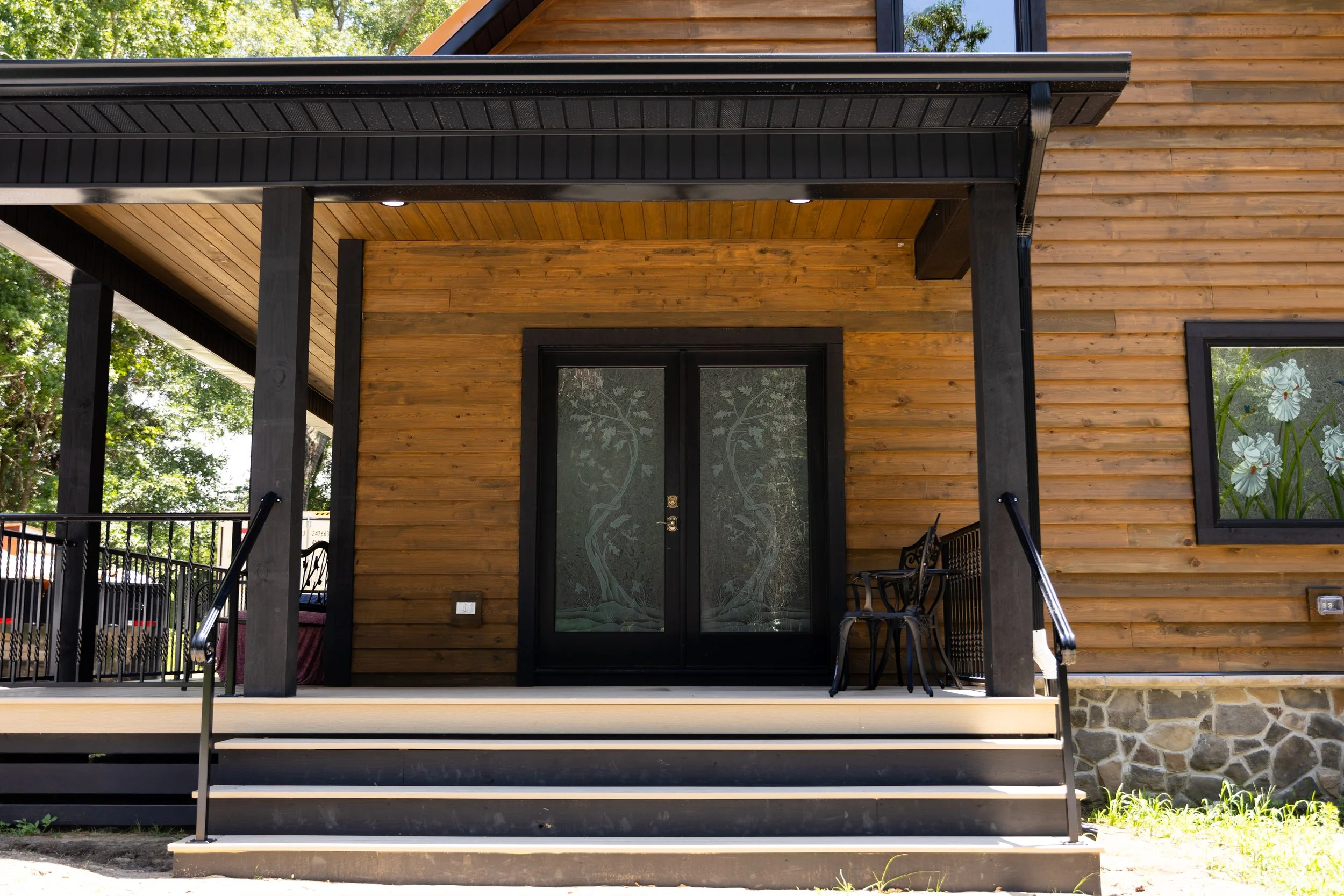 Front view of a modern house with a wooden exterior, black framed glass doors with tree design, and a small porch with black metal railings and steps.