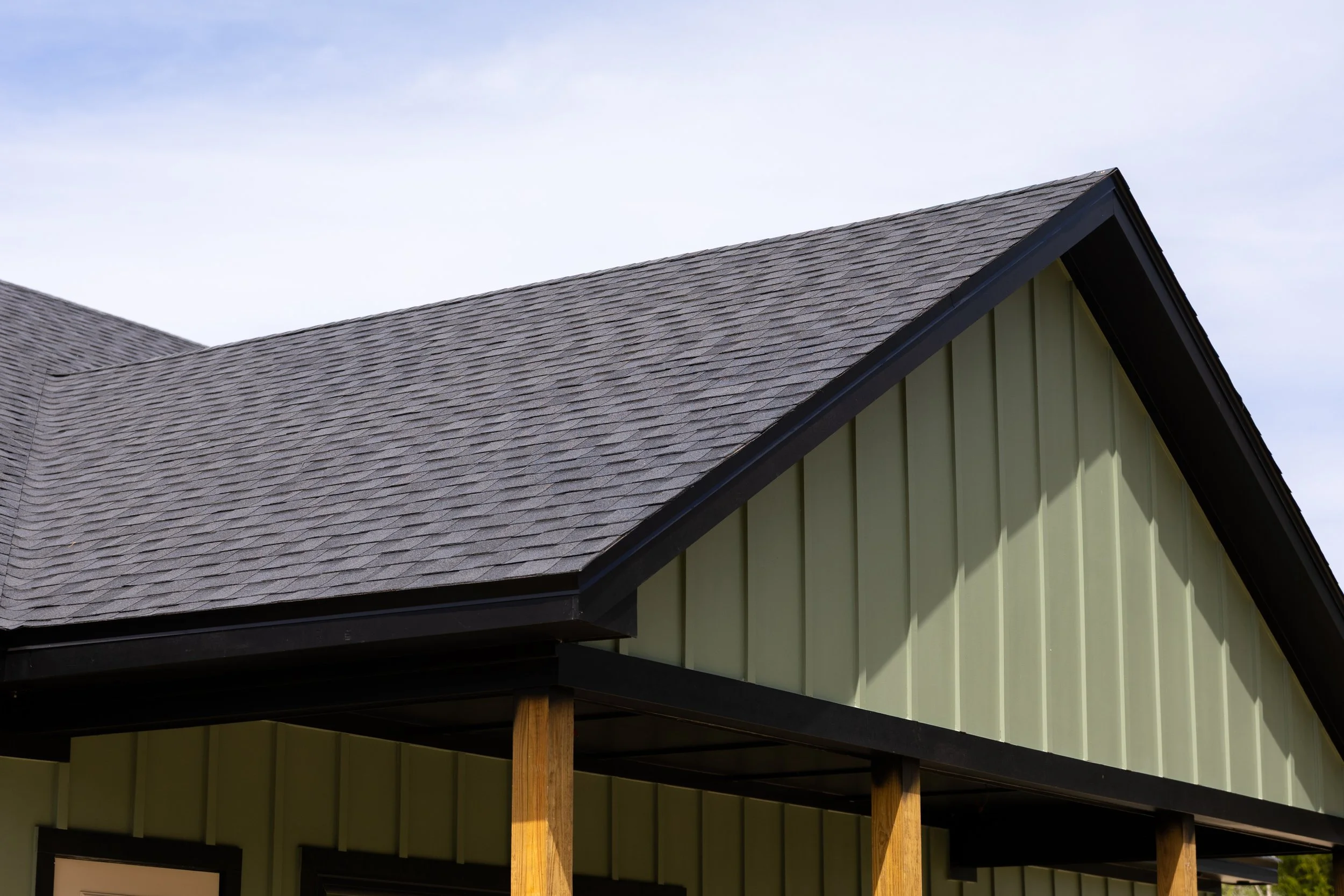 The image shows the roof of a house with dark gray shingles, extending over a porch area supported by wooden beams. The siding of the house is light green with vertical panels. The sky is partly cloudy, with blue and white clouds visible.