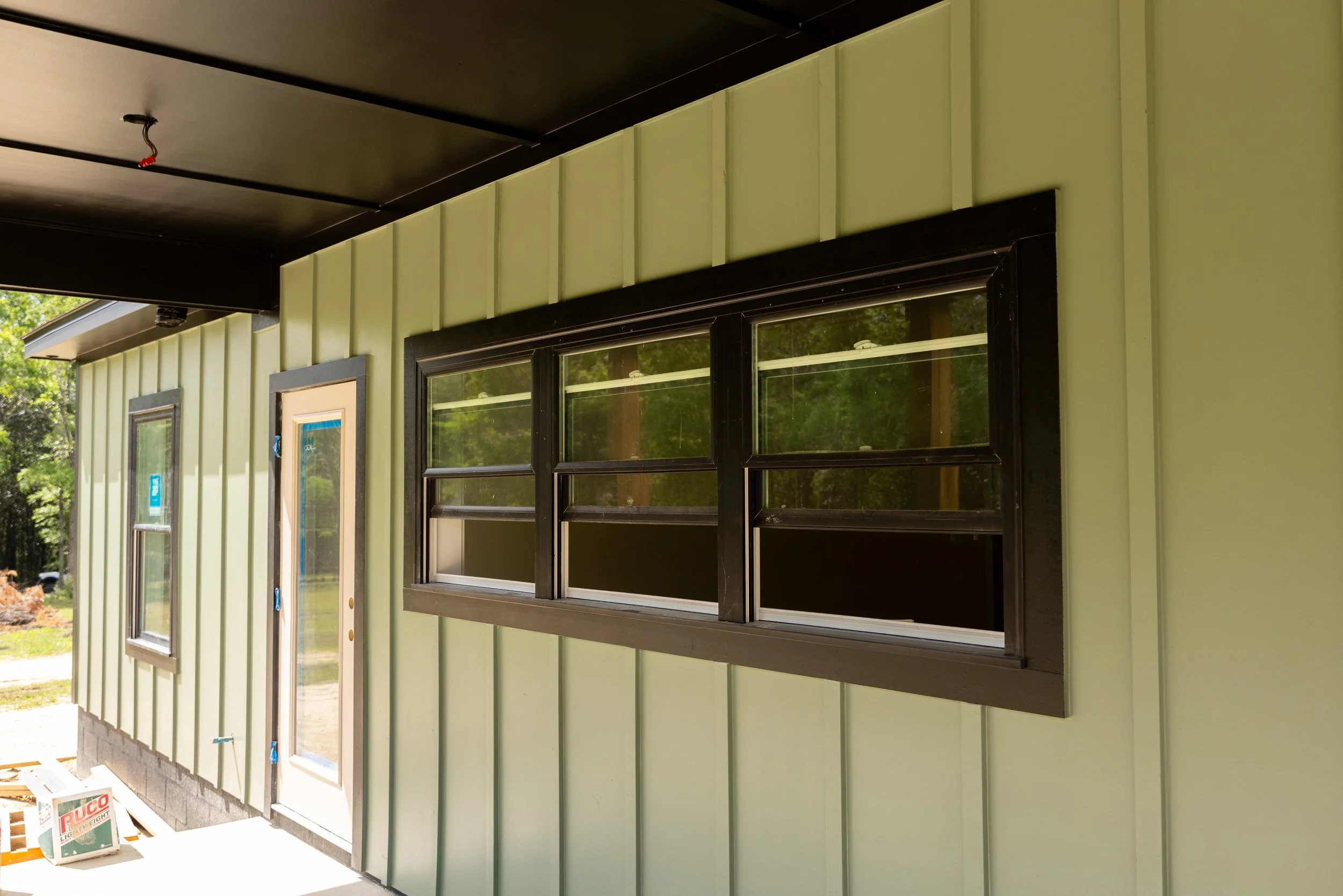 Exterior of a house under construction with green siding, black window frames, and a black soffit, with a small window and door on the left side, and construction materials on the ground.