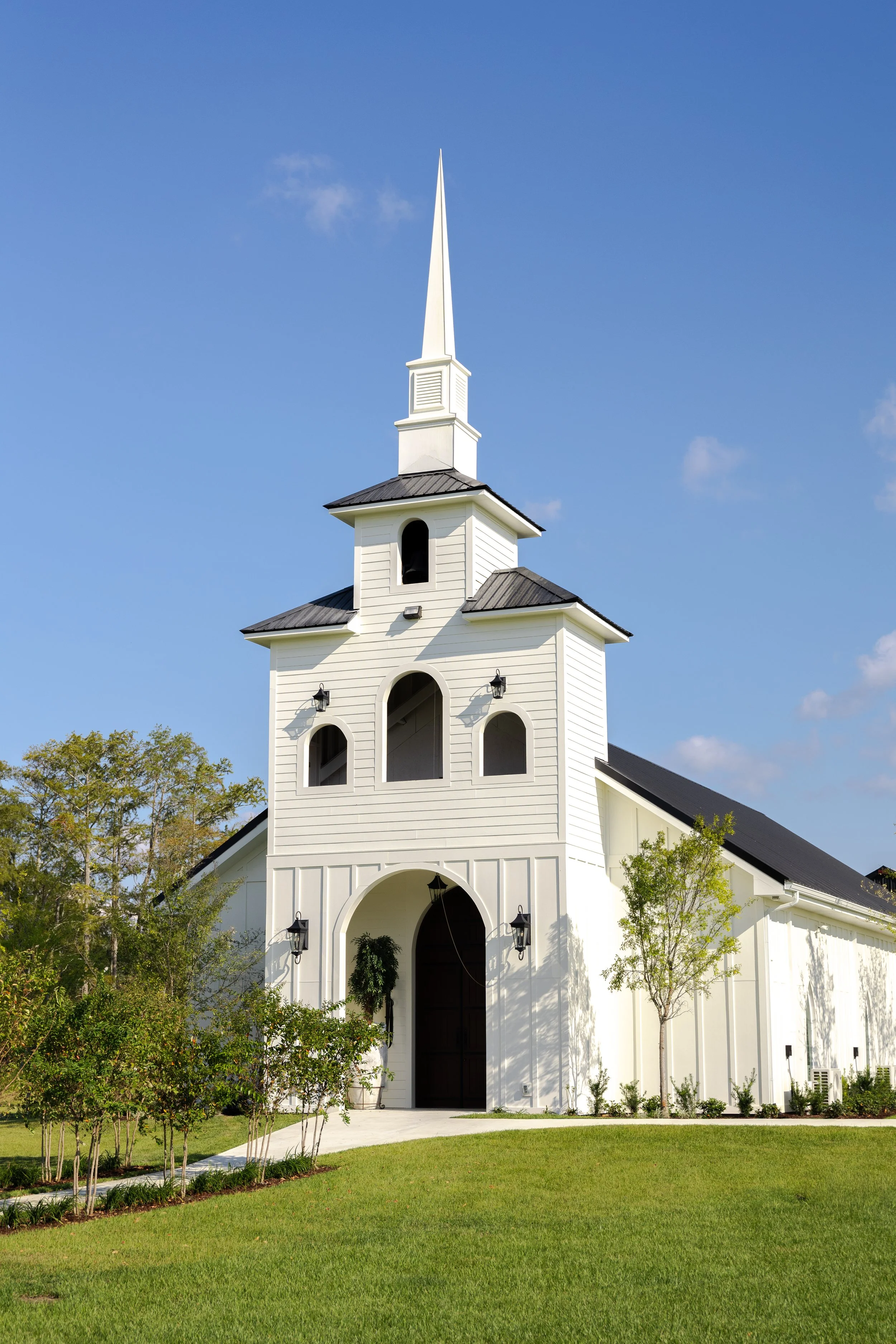 White church with black roof and tall steeple, set against a blue sky, surrounded by green trees and grass.