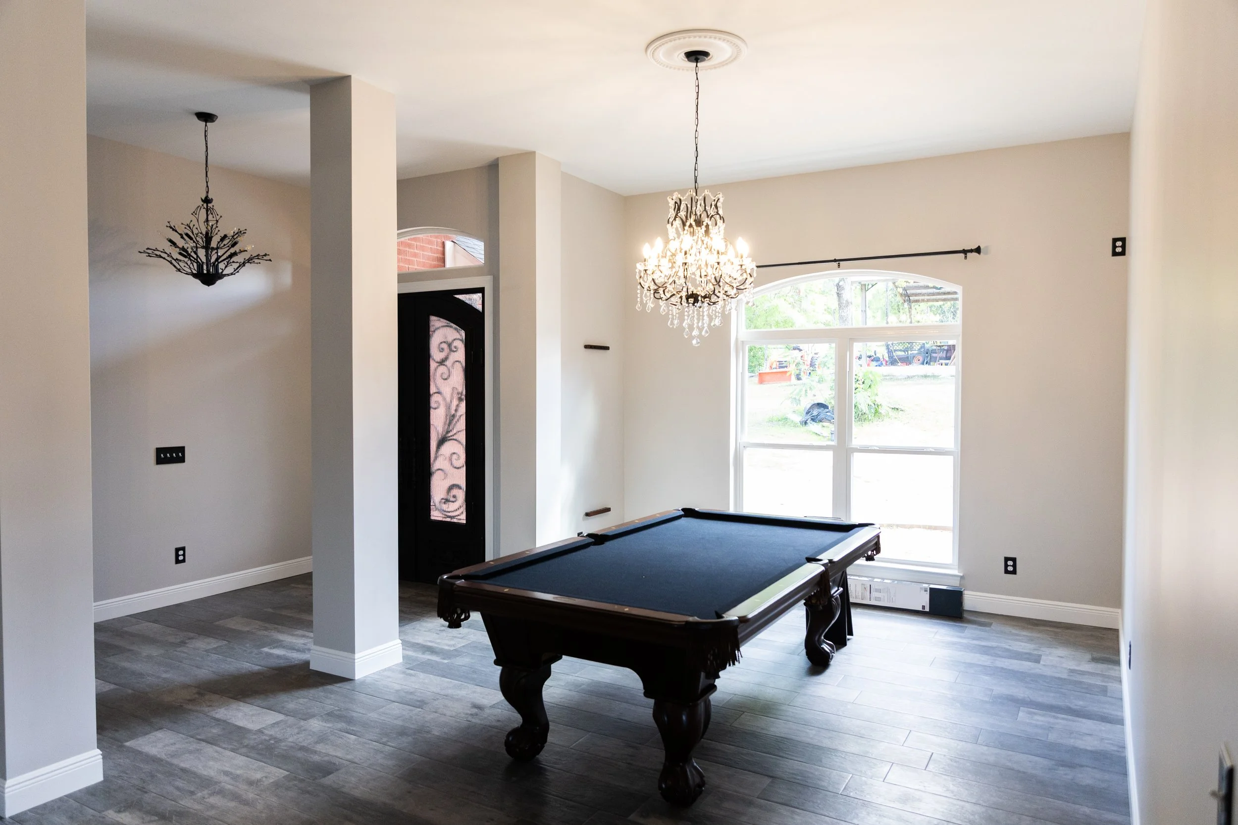Living room with a black pool table, a chandelier hanging from the ceiling, a large window letting in natural light, and an ornate front door with decorative glass design.
