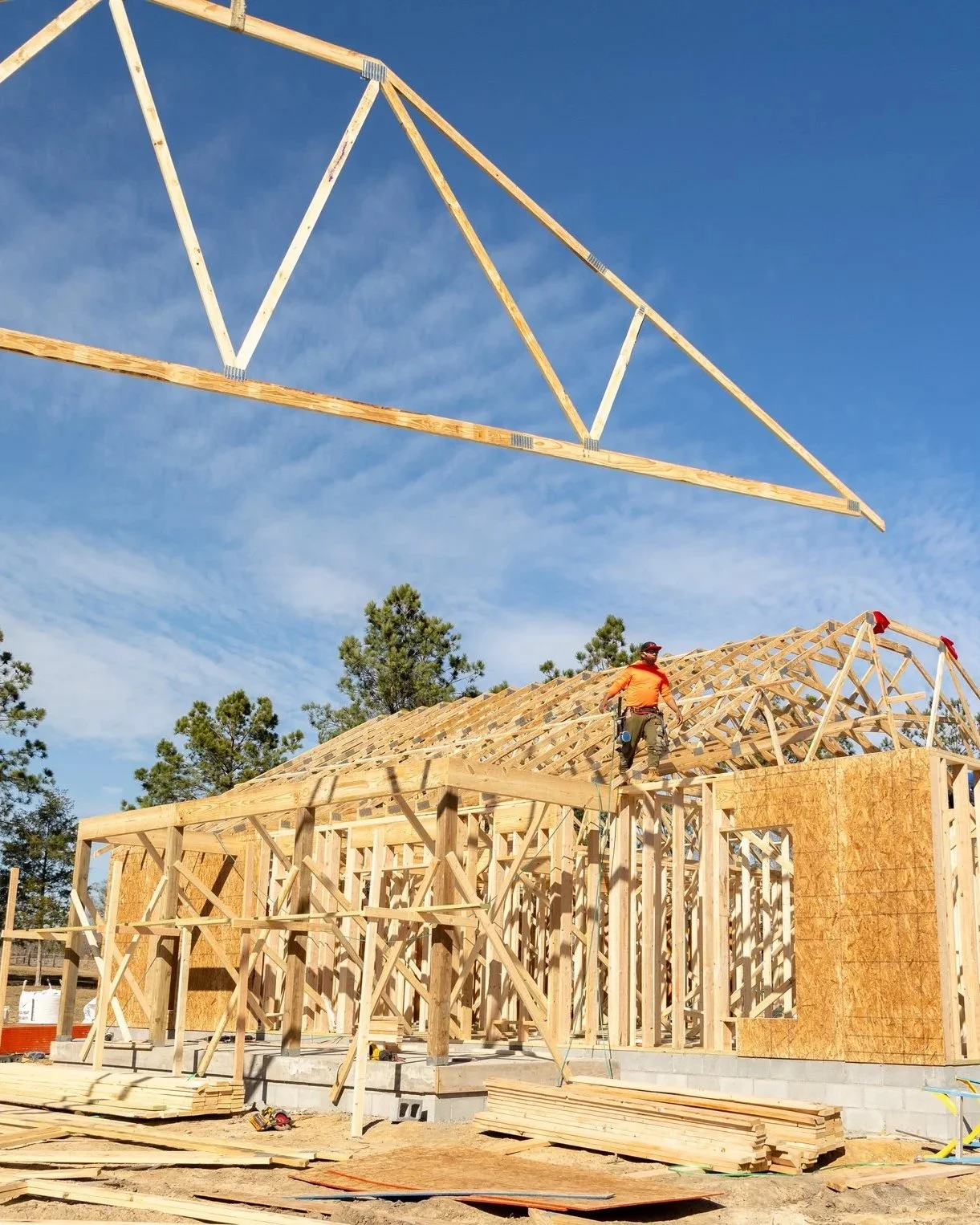 A construction worker wearing an orange shirt and a safety helmet stands on the wooden framework of a house under construction, with wooden beams and a roof structure being built, and a crane lifting a large wooden truss above the building against a clear blue sky.