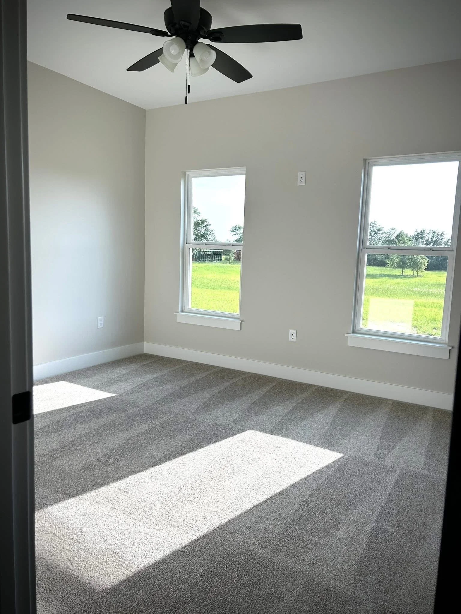 An empty room with gray walls, a ceiling fan with lights, and three windows showing a grassy landscape outside, sunlight casting shadows on the carpeted floor.