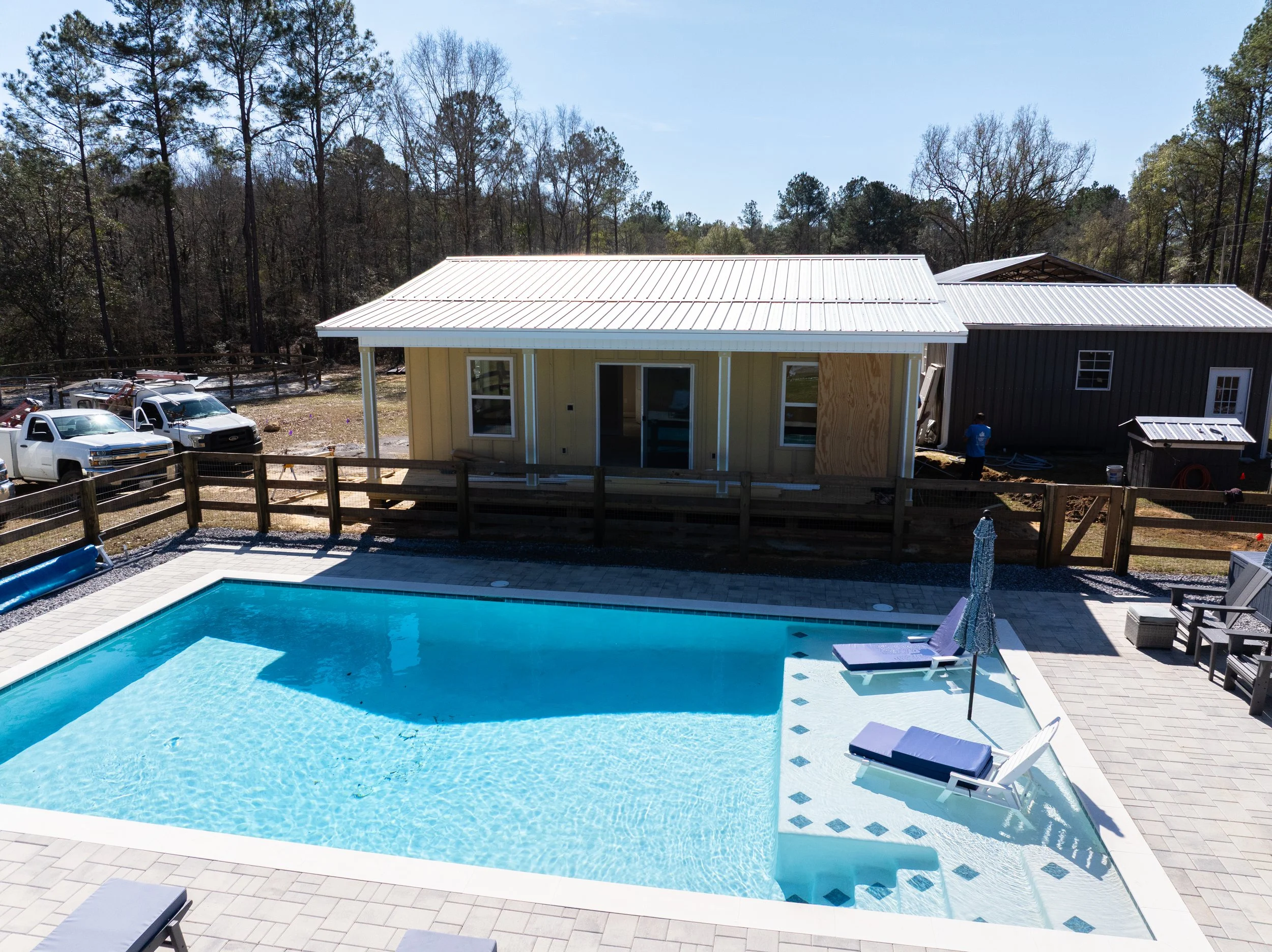 A backyard with a swimming pool, poolside chairs, and a fenced area. There are two houses under construction, one painted yellow with a metal roof, and another partially visible with dark siding. Several trucks are parked nearby, and a person is work