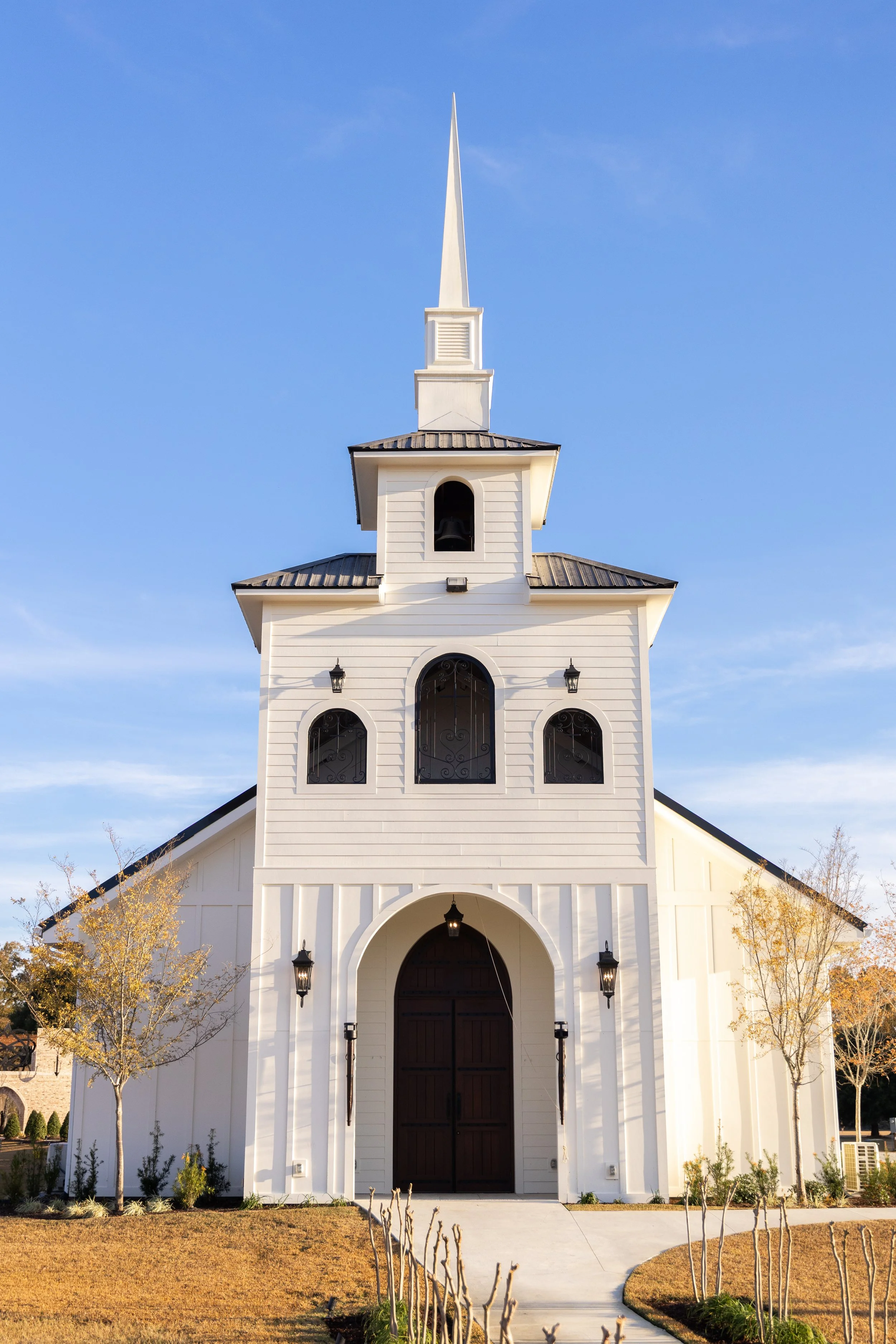 A white church with a tall steeple, arched windows, and a dark wooden door, surrounded by a landscaped yard with trees and a pathway.