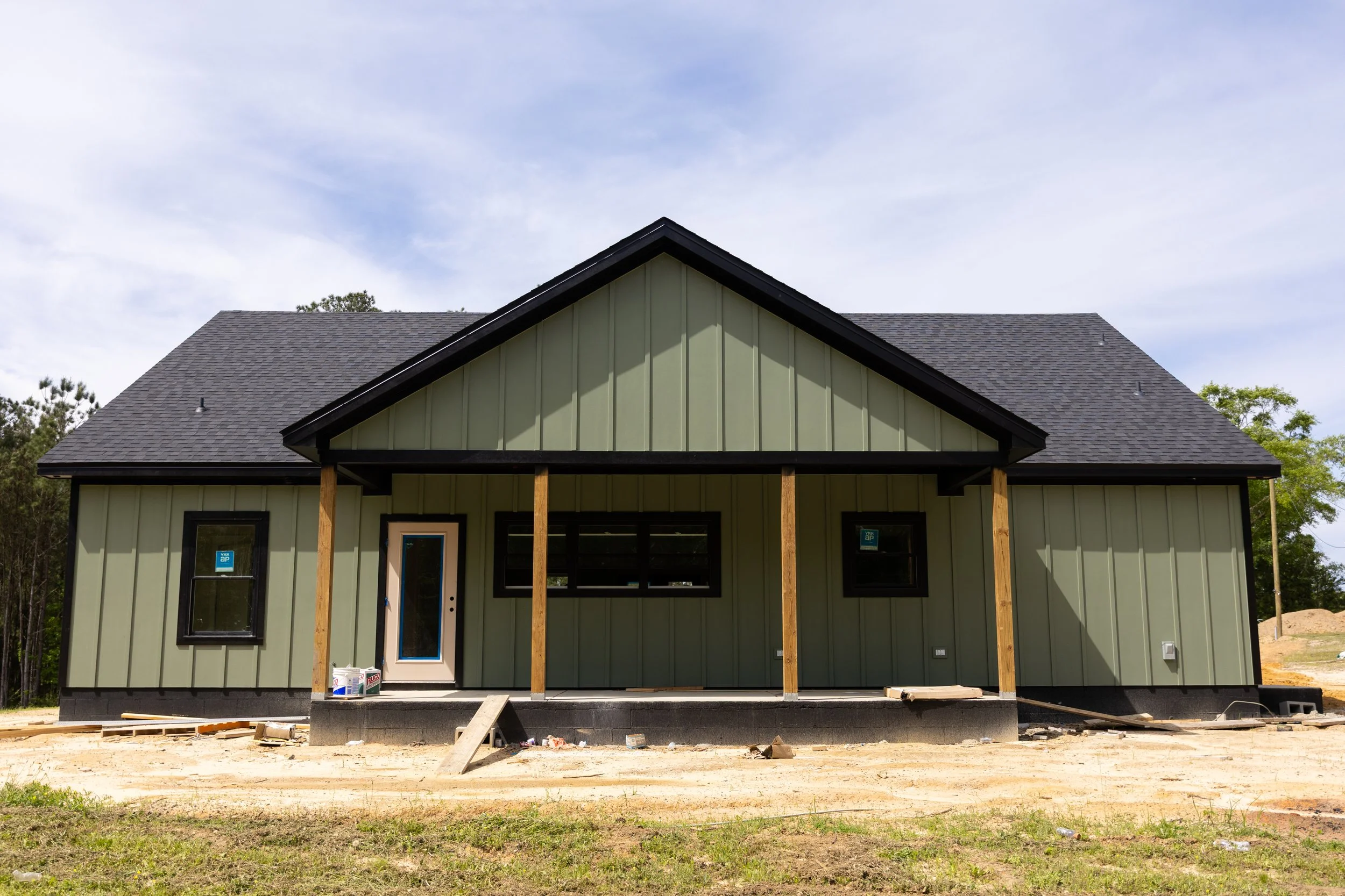 Front view of a house under construction with dark gray roof and light green vertical siding, with wooden support beams and construction materials in front.