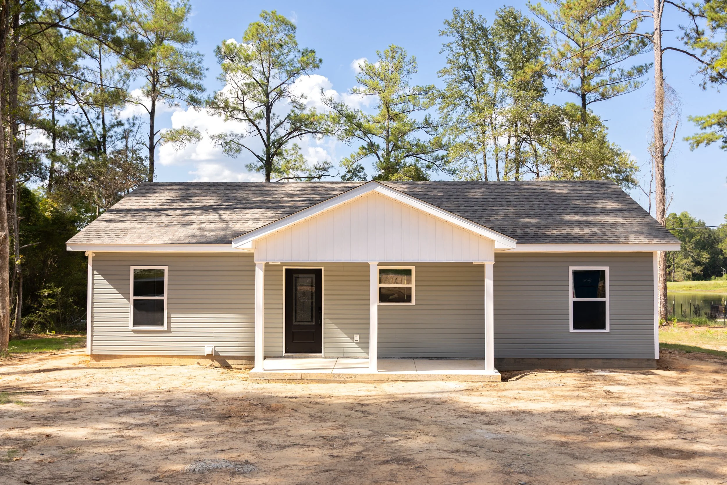 Front view of a house under construction with a new porch, gray siding, and a black front door, surrounded by tall trees and a partly cloudy sky.