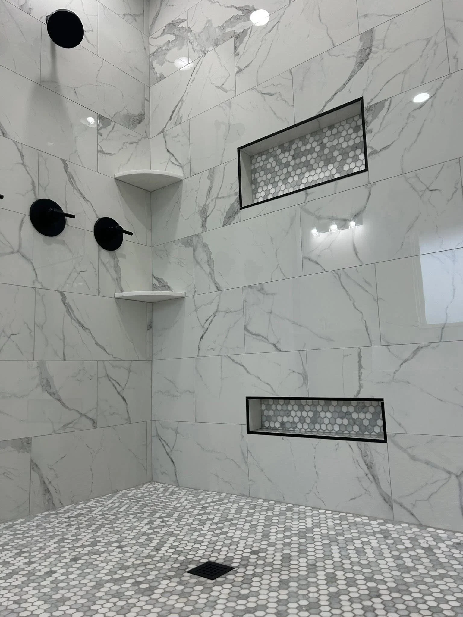 A modern walk-in shower with marble wall tiles, two black shower controls, a small corner shelf, two built-in niches with hexagonal tile backs, and a circular white and gray hexagonal tiled floor with a black drain in the center.
