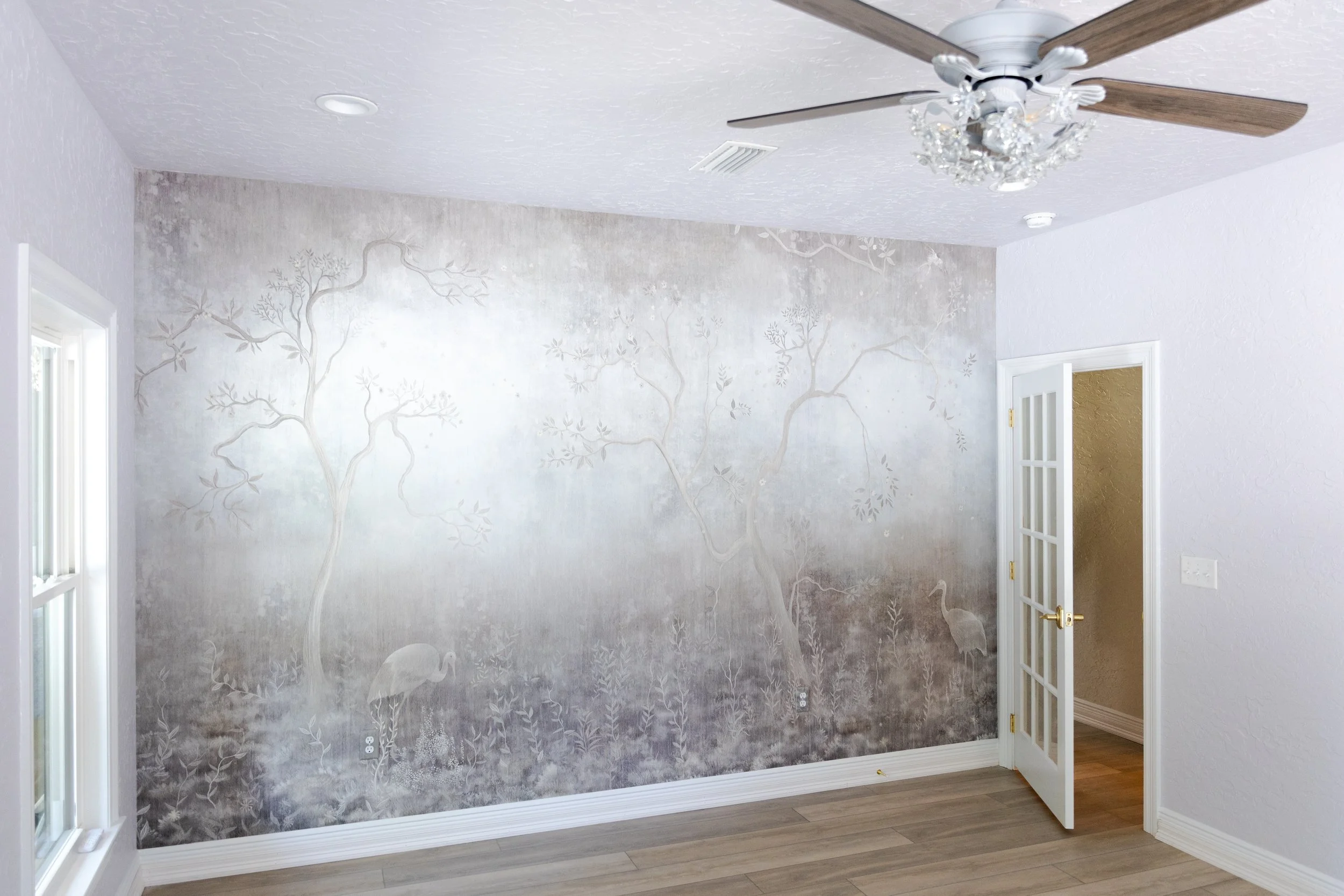A room with a nature-themed mural of trees and flamingos on one wall, a ceiling fan with crystal accents, a window on the left, and an open door leading to another room.