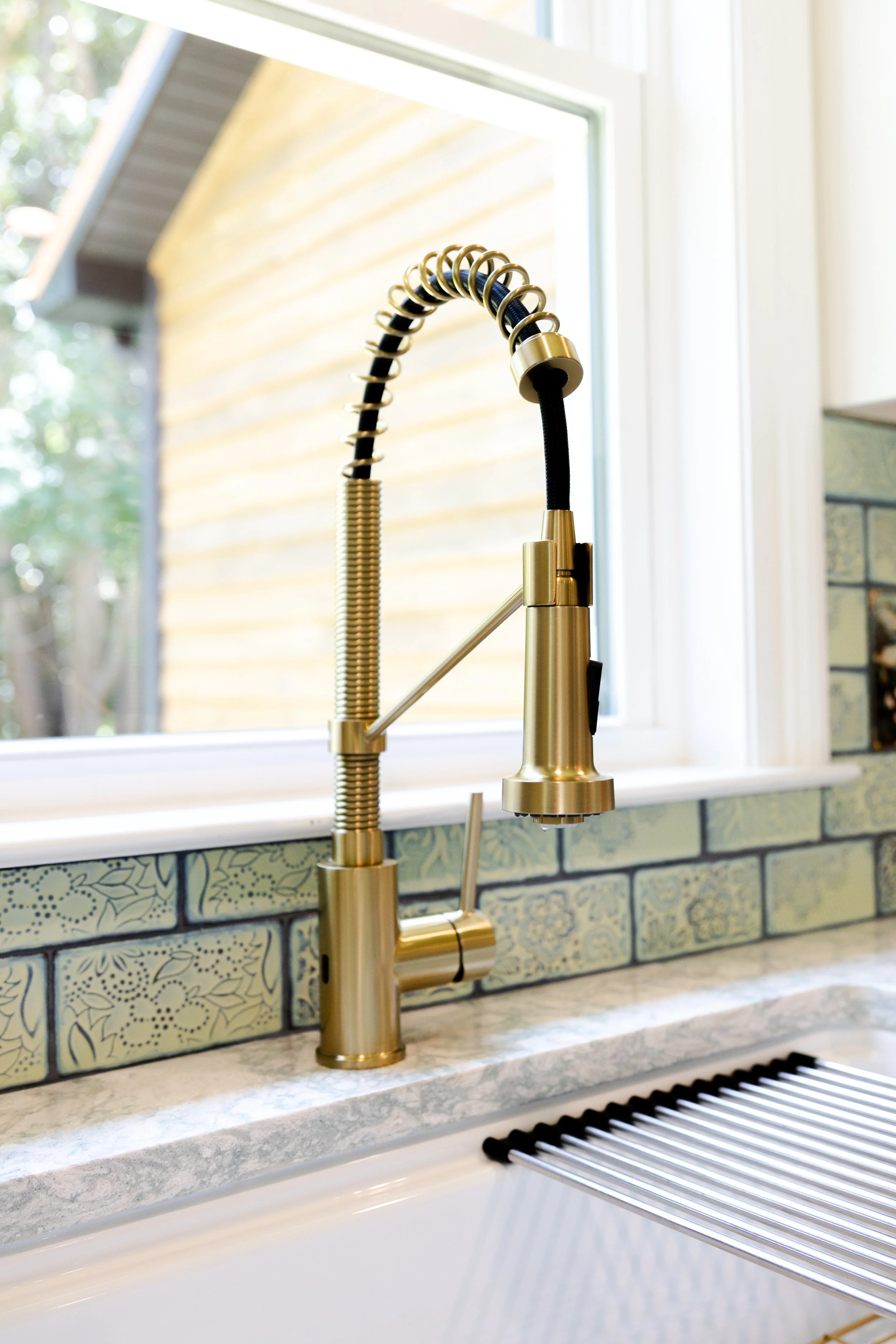 Close-up of a modern kitchen faucet with a high-arc spout and spring coil design, set against a window and decorative tile backsplash.