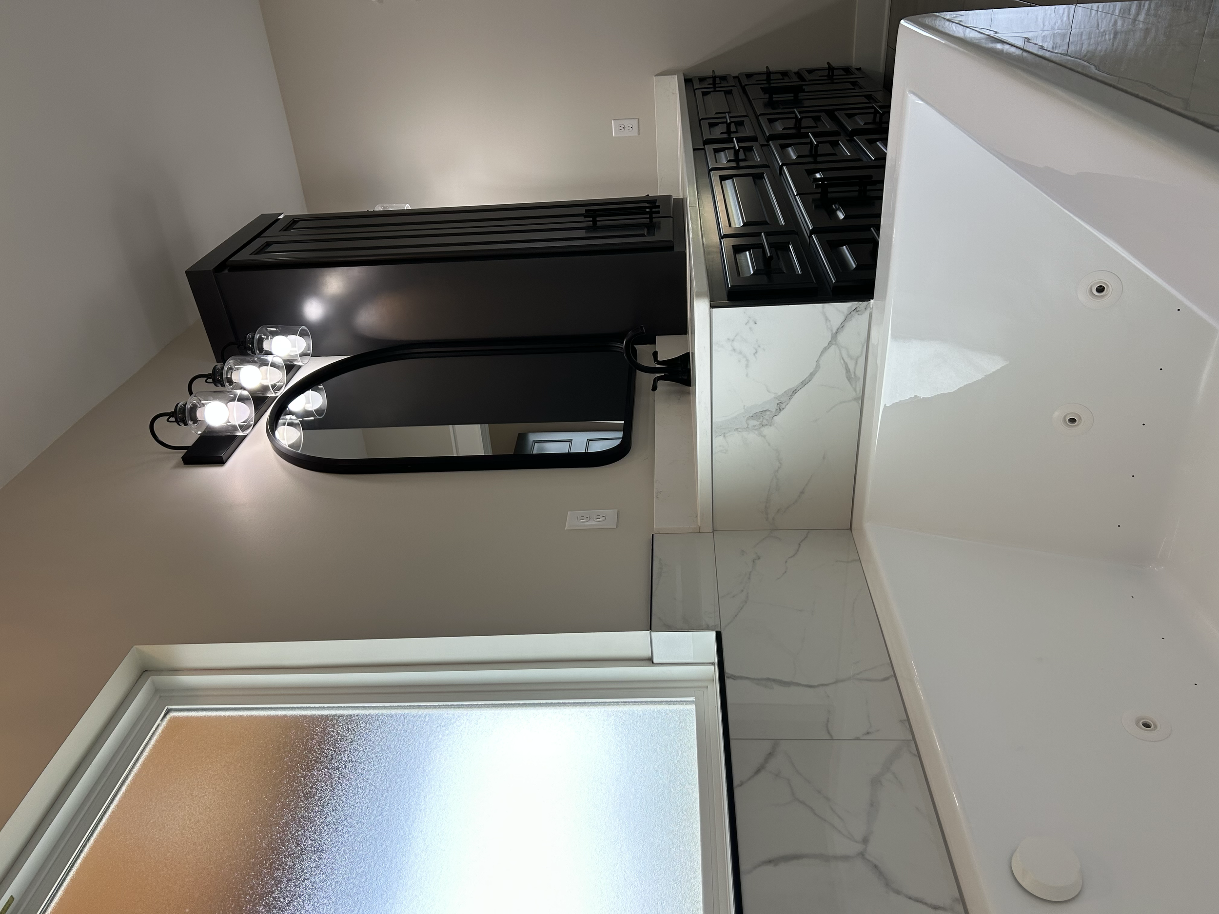 Modern kitchen with black cabinets, white marble countertop, and a black stove. A mirror with vanity lights is visible, and a frosted window with natural light is in the background.