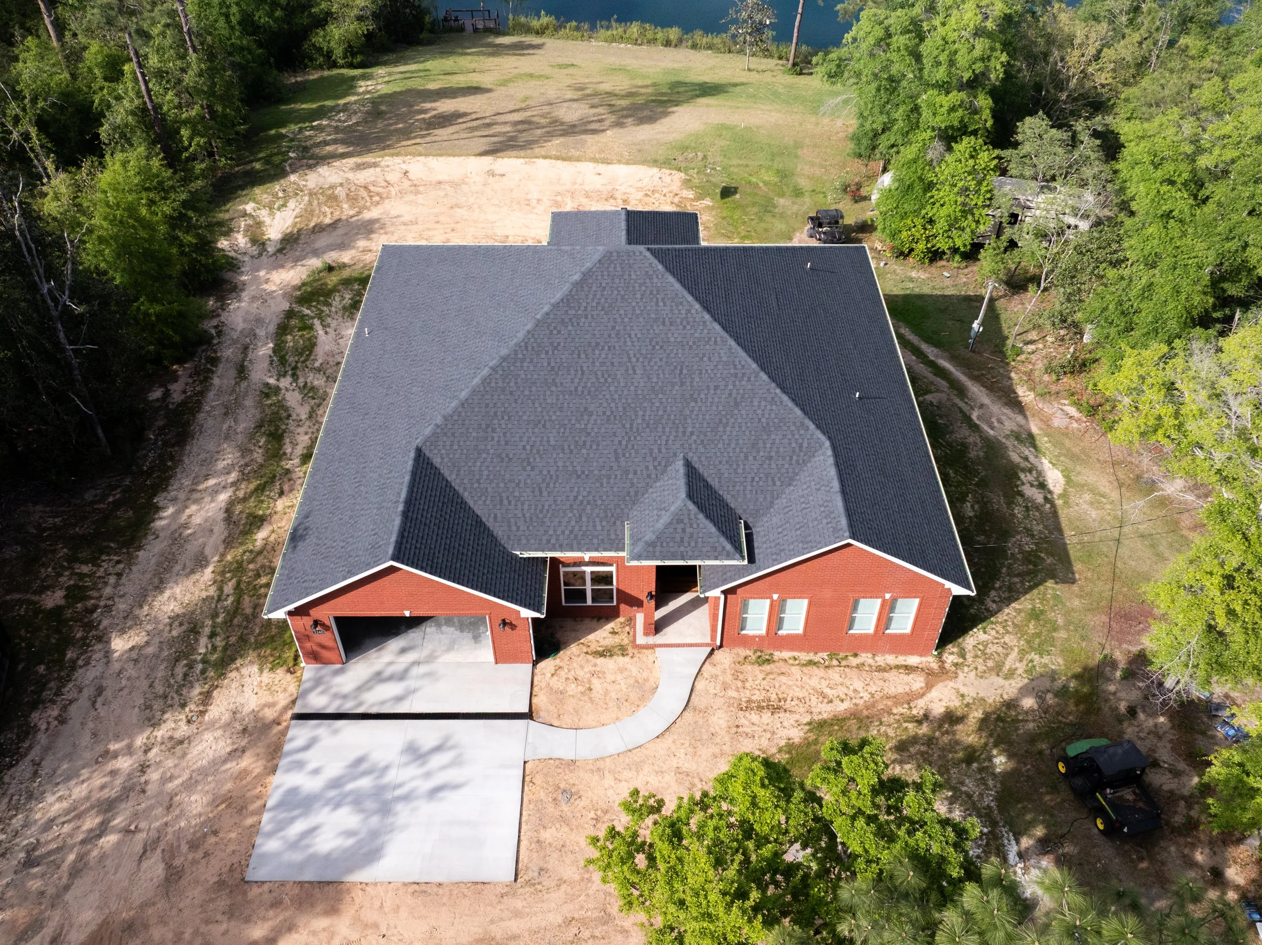 Aerial view of a house with a gray roof and brick exterior, surrounded by trees and a driveway leading to a garage.