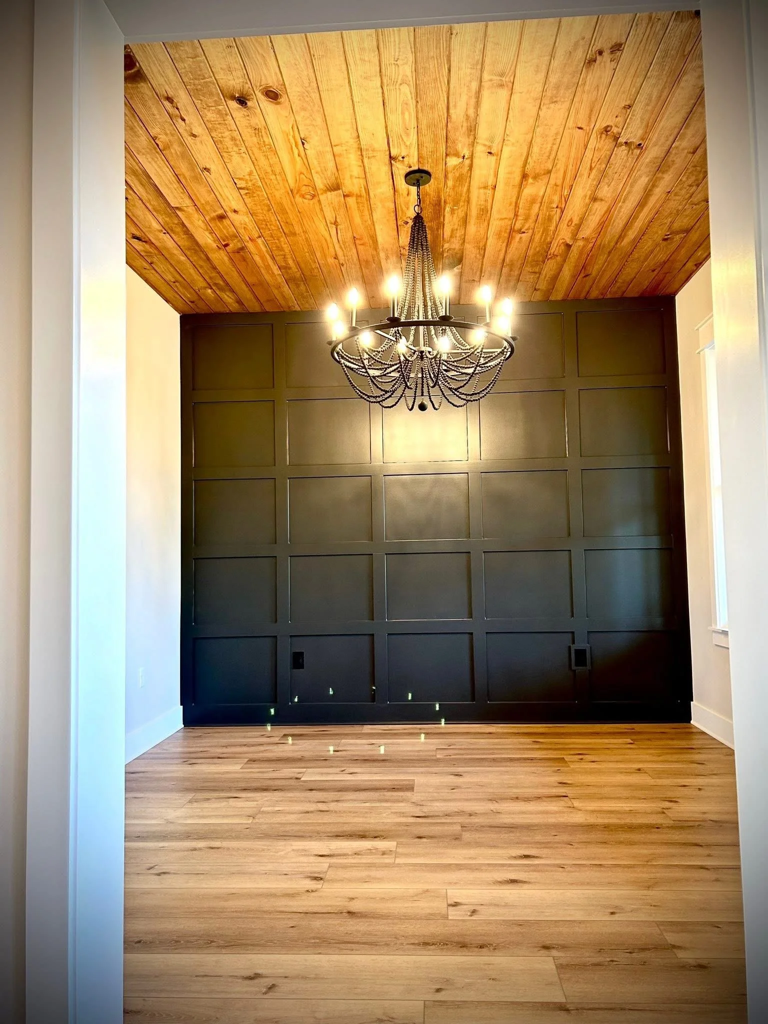 An interior view of a room with natural wood flooring, a feature wall with black paneling, and a wooden ceiling. A black chandelier with lit bulbs hangs from the ceiling.