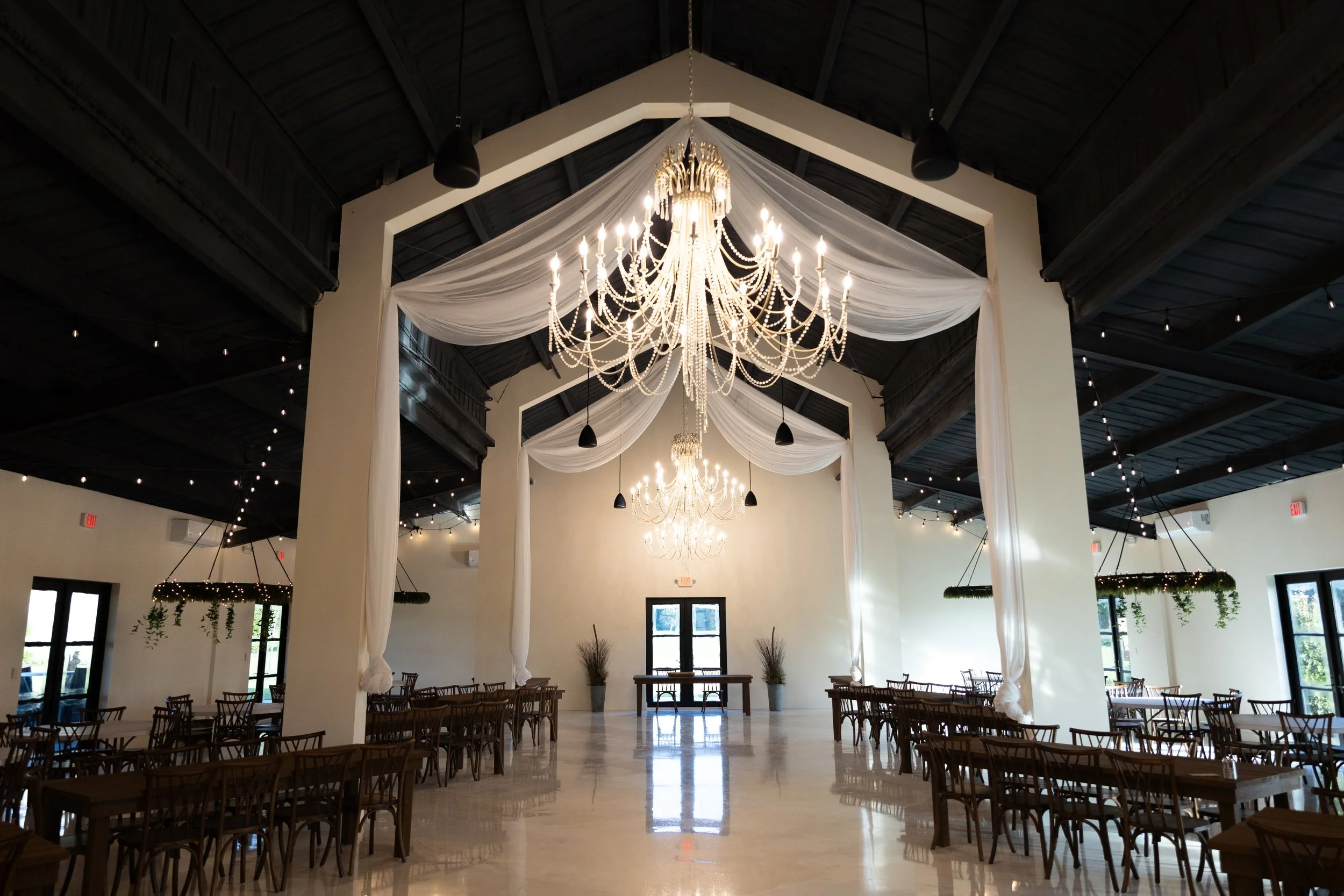 Interior of an event space with a large chandelier, draped fabric decorations, dark wood ceiling, and multiple tables and chairs.