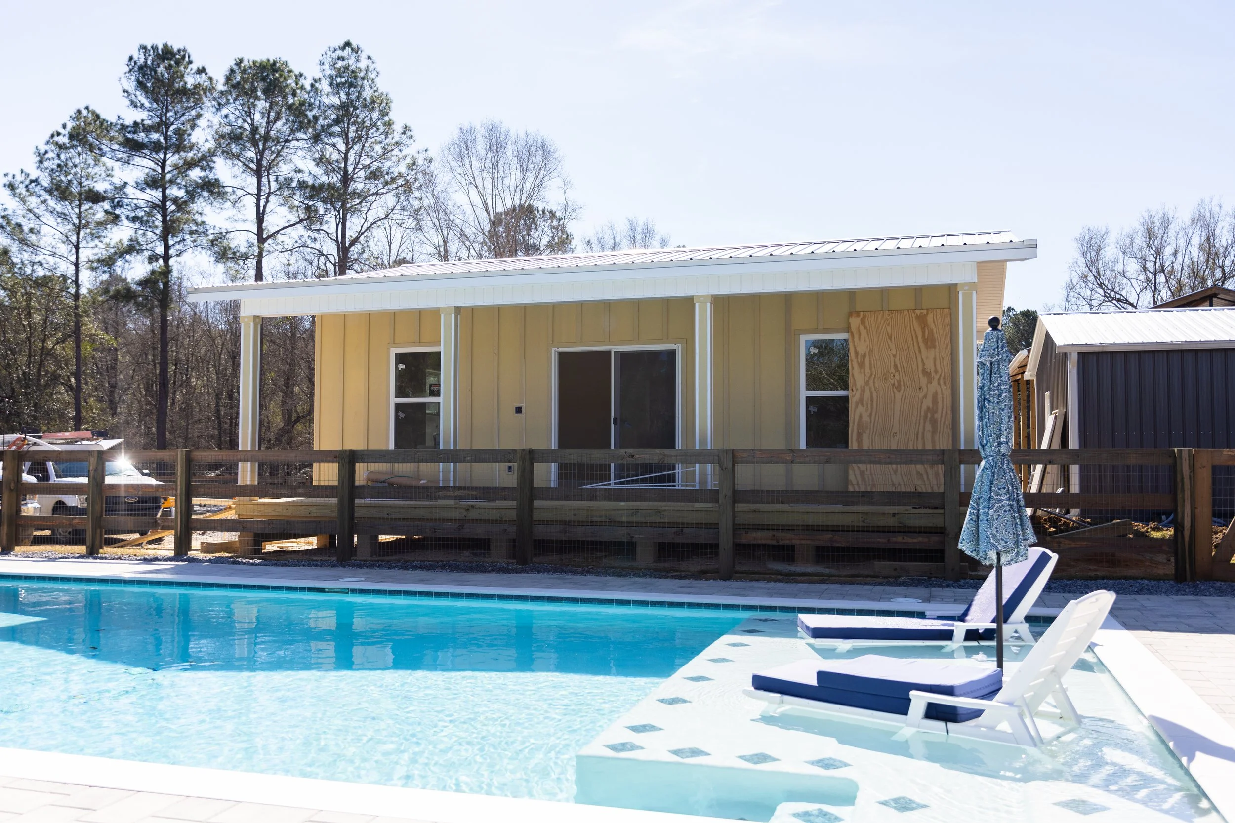 Swimming pool with lounge chairs and a blue umbrella, house under construction in the background, trees and clear sky.