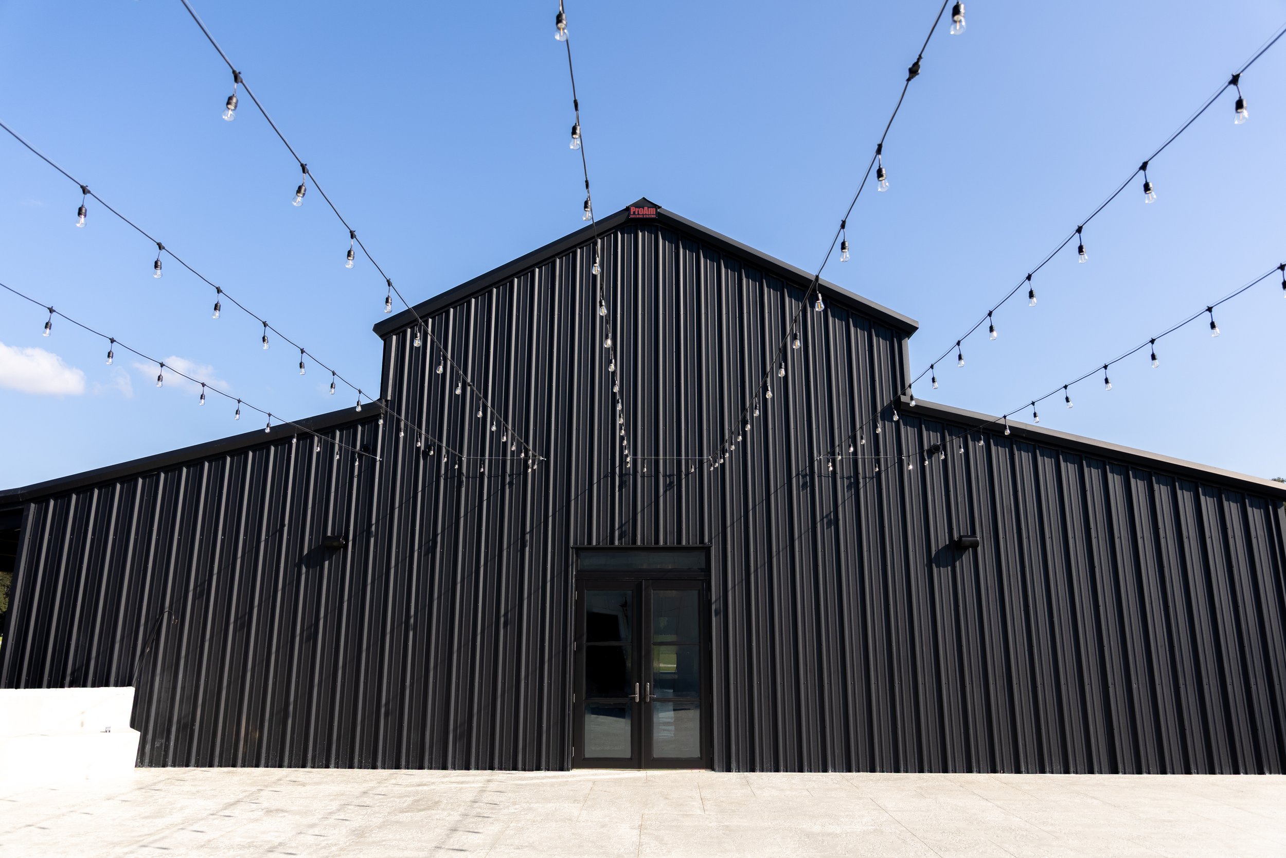 Black modern building with vertical metal siding, glass doors, and string lights overhead against a blue sky.