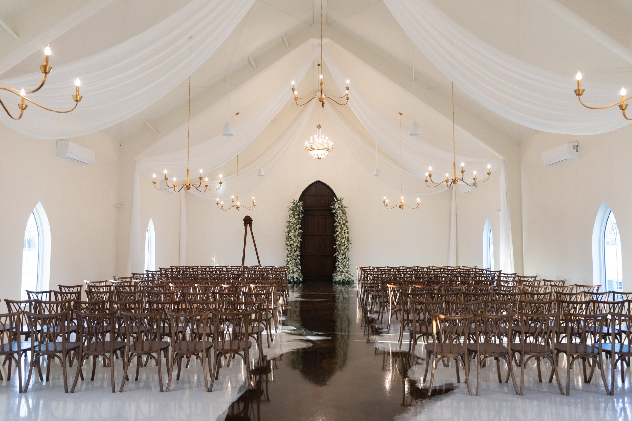 A decorated indoor wedding ceremony venue with rows of wooden chairs facing an altar adorned with white flowers, elegant drapery hanging from the ceiling, and chandeliers.