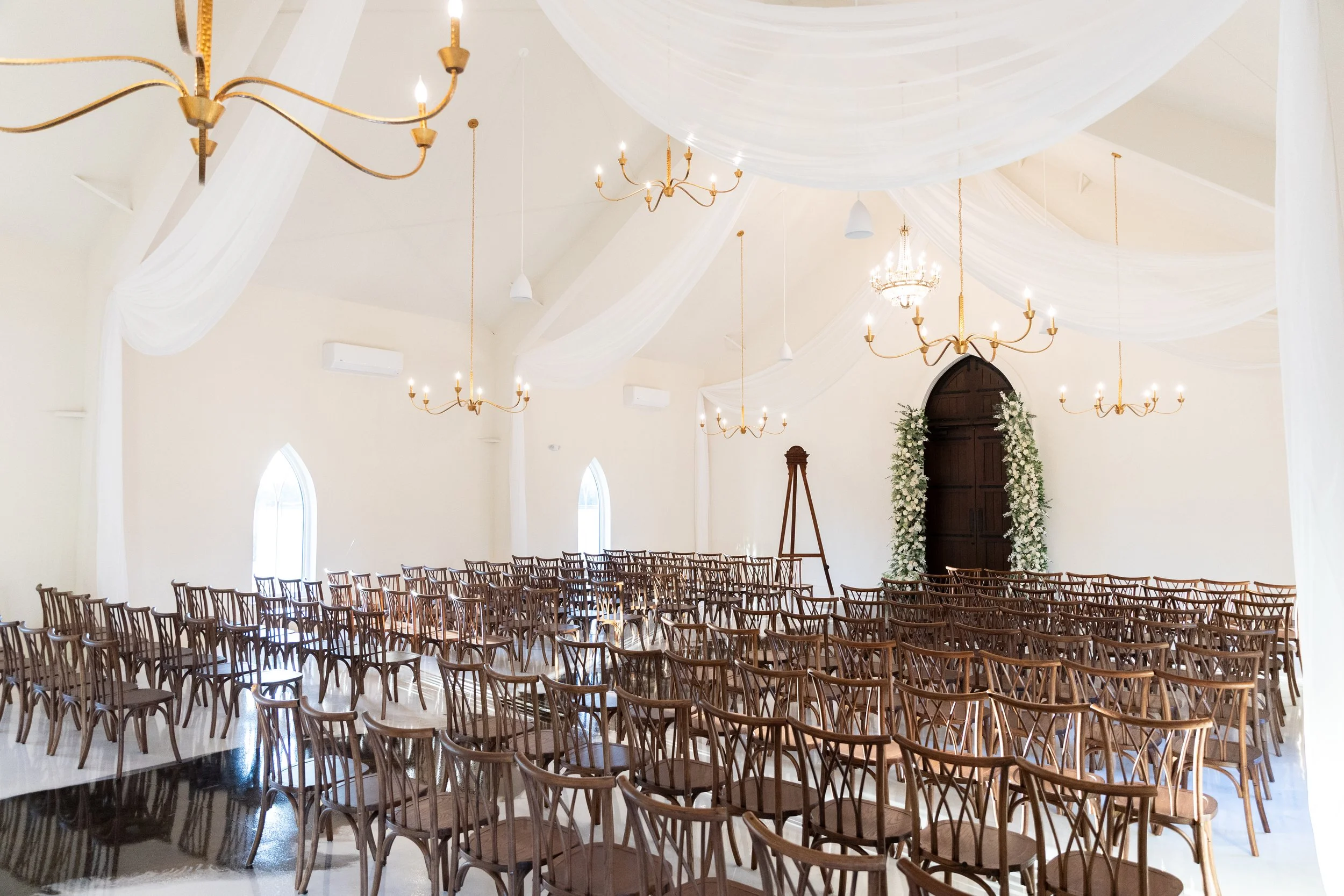Interior of a church decorated for a wedding with rows of wooden chairs, chandeliers, and draped white fabric ceiling, with floral arch at the front.