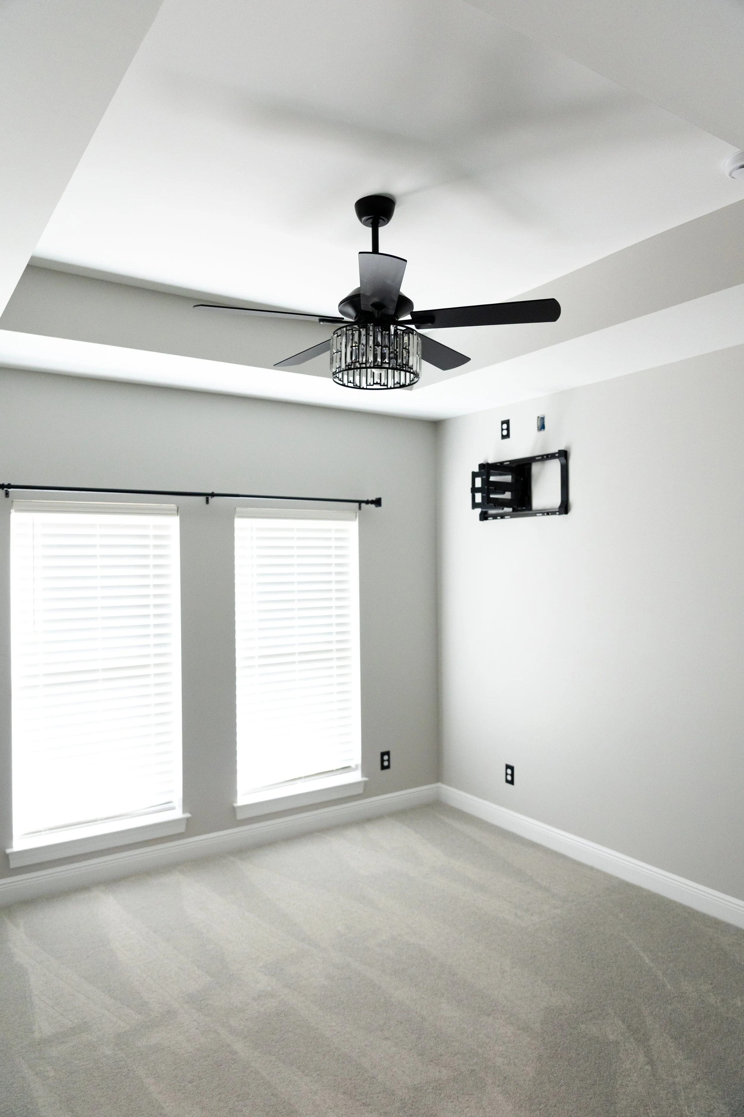 Empty room with two windows covered with white blinds, a ceiling fan with a light fixture, and a wall mount for a television or monitor.