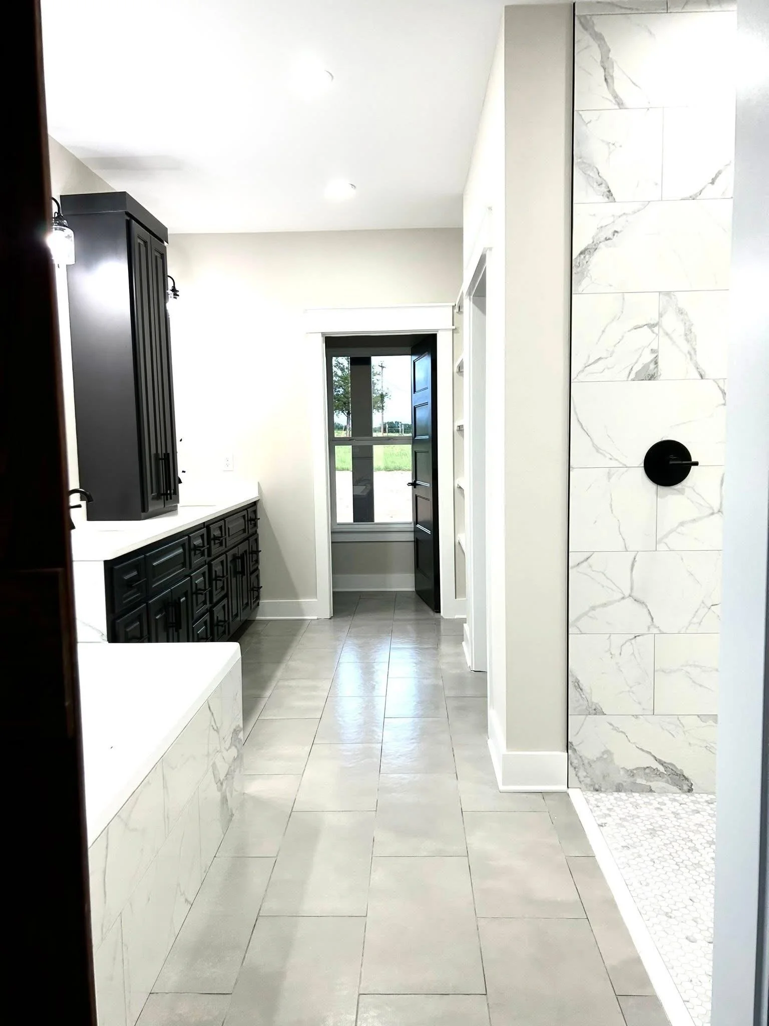 Modern bathroom with white marble walls, gray floor tiles, black cabinets, and a small shower area with hexagonal tiles.