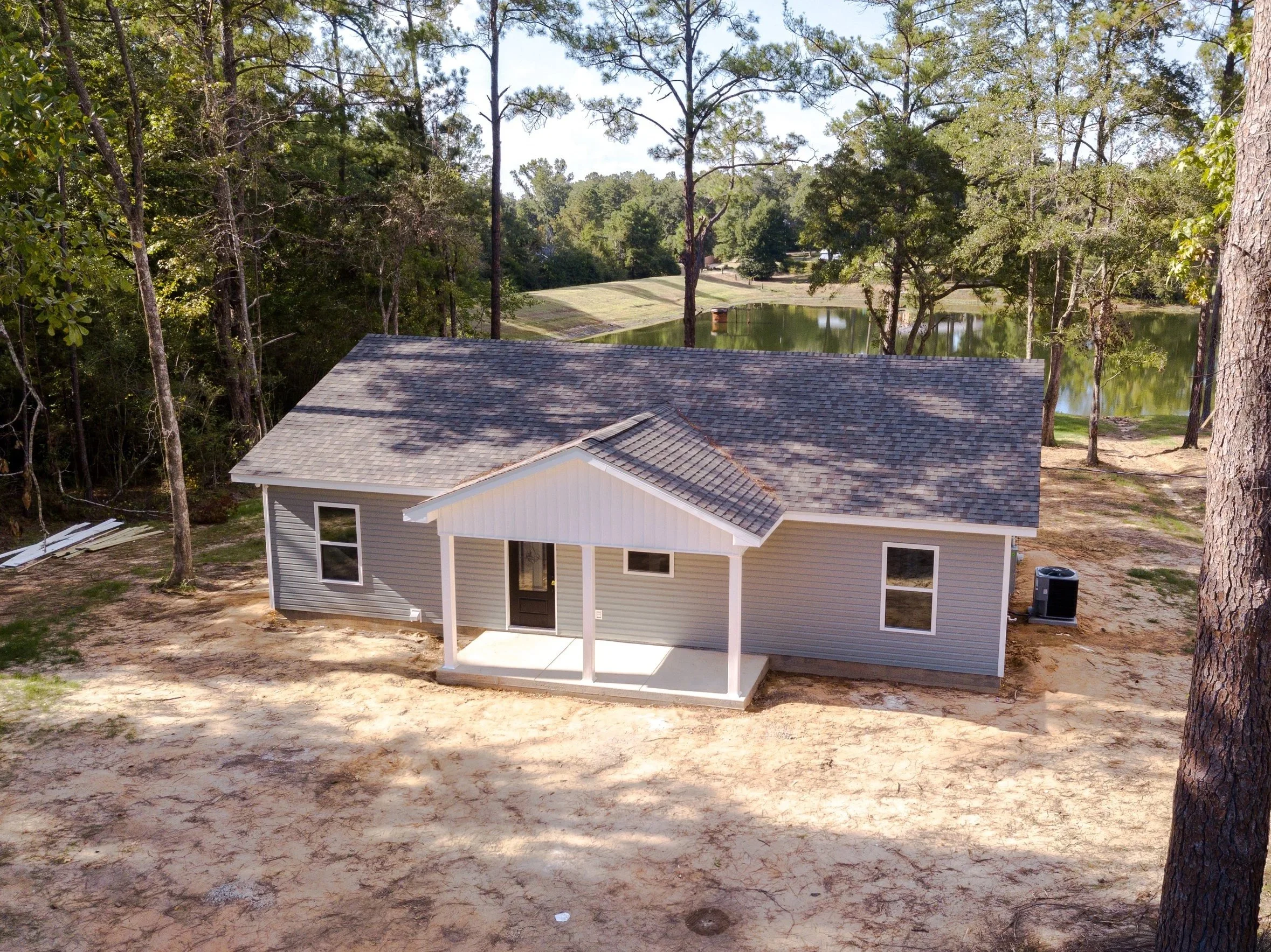A house with gray siding and a small covered front porch, located in a wooded area near a pond, with tall trees surrounding it and a dirt yard in the foreground.