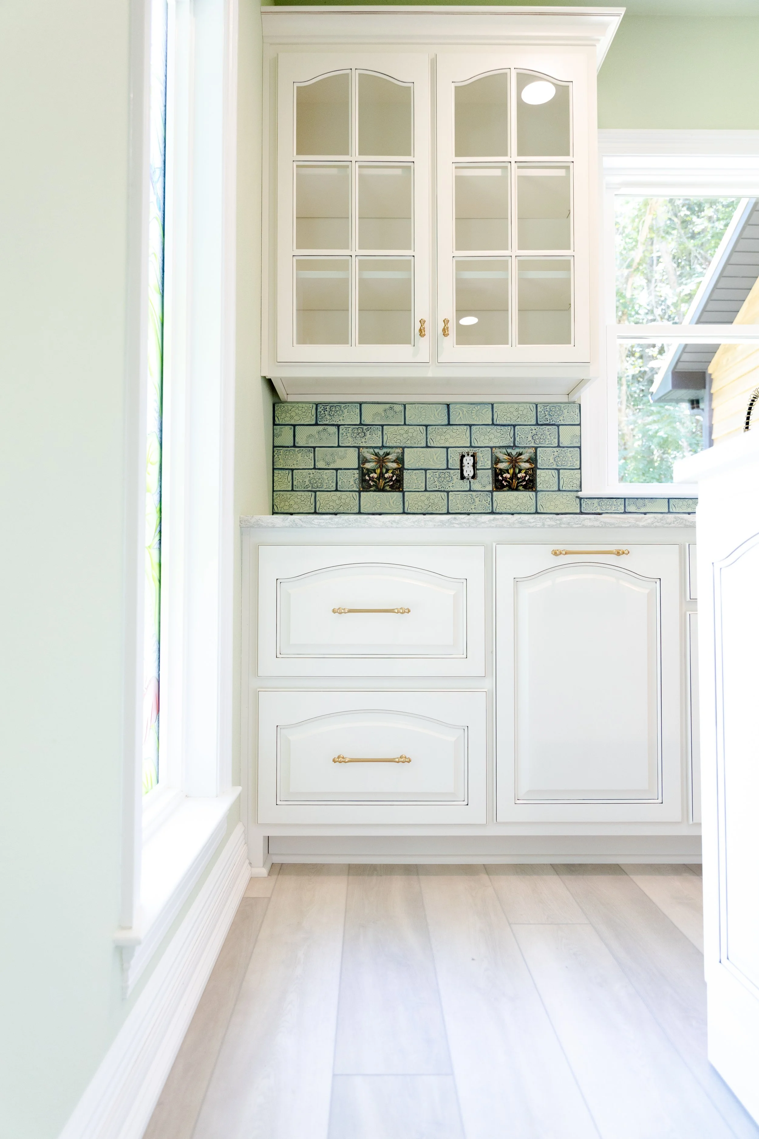 view of a kitchen or built-in cabinet area with white cabinets, a decorative tile backsplash featuring bees and flowers, a window on the right side, light-colored floor tiles, and a green wall.